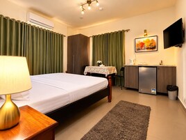 Habitación de hotel con cama, escritorio con lámpara, mesita con un jarrón, televisor y una pequeña cocina.