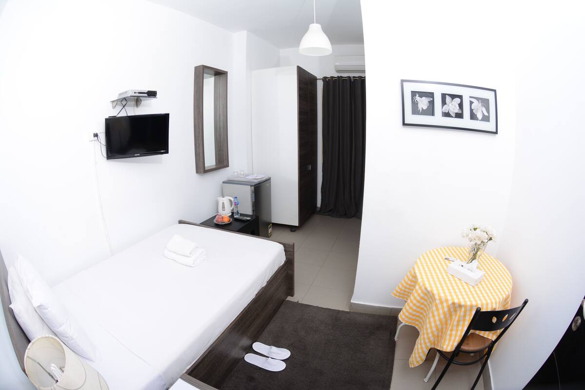 A hotel room with a bed, a desk with a chair, a TV, a mirror, and a small table with a vase of flowers.