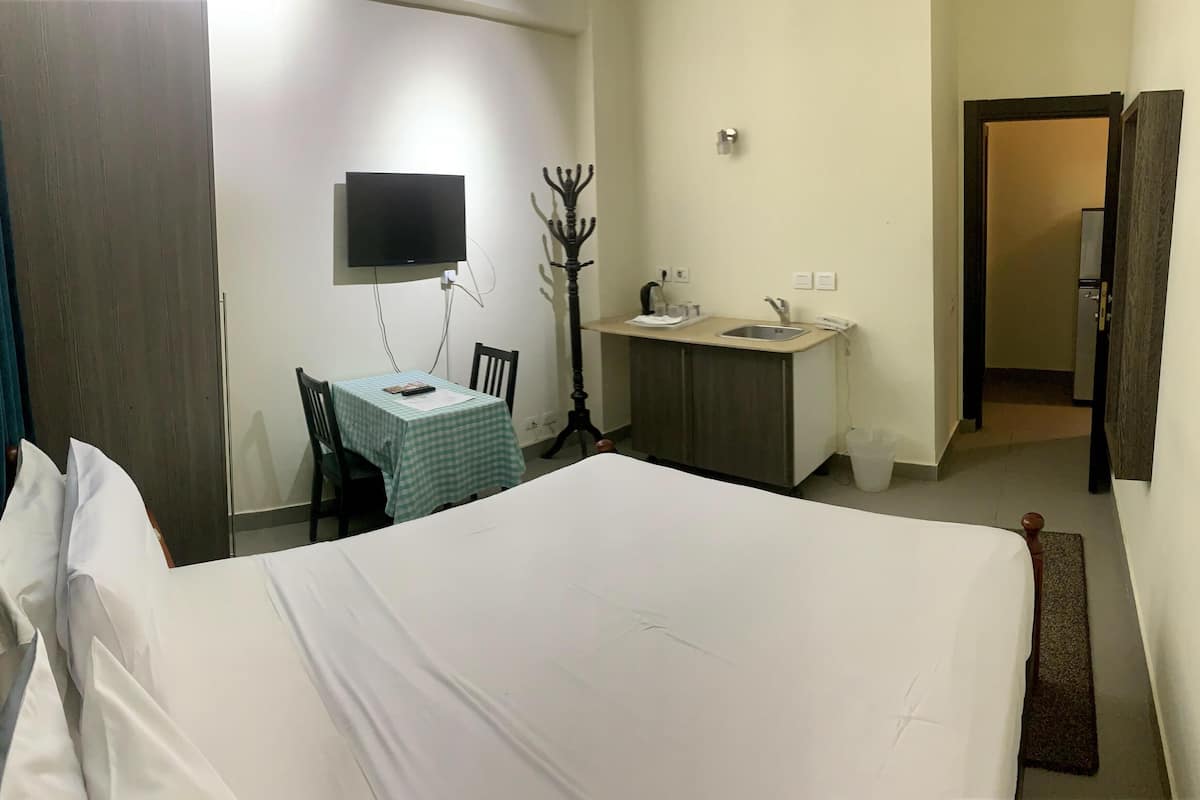 A hotel room with a bed, a desk with a chair, a TV mounted on the wall, and a small kitchenette area.