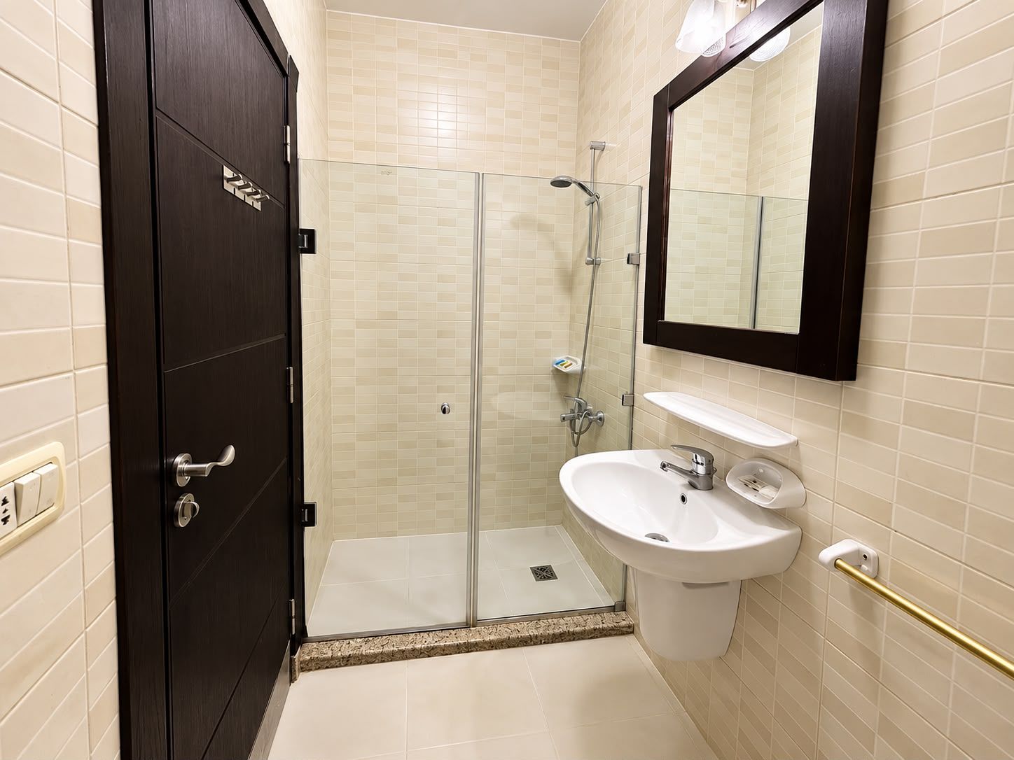 A bathroom with a glass-enclosed shower, a white sink, a mirror, and a dark wooden door.