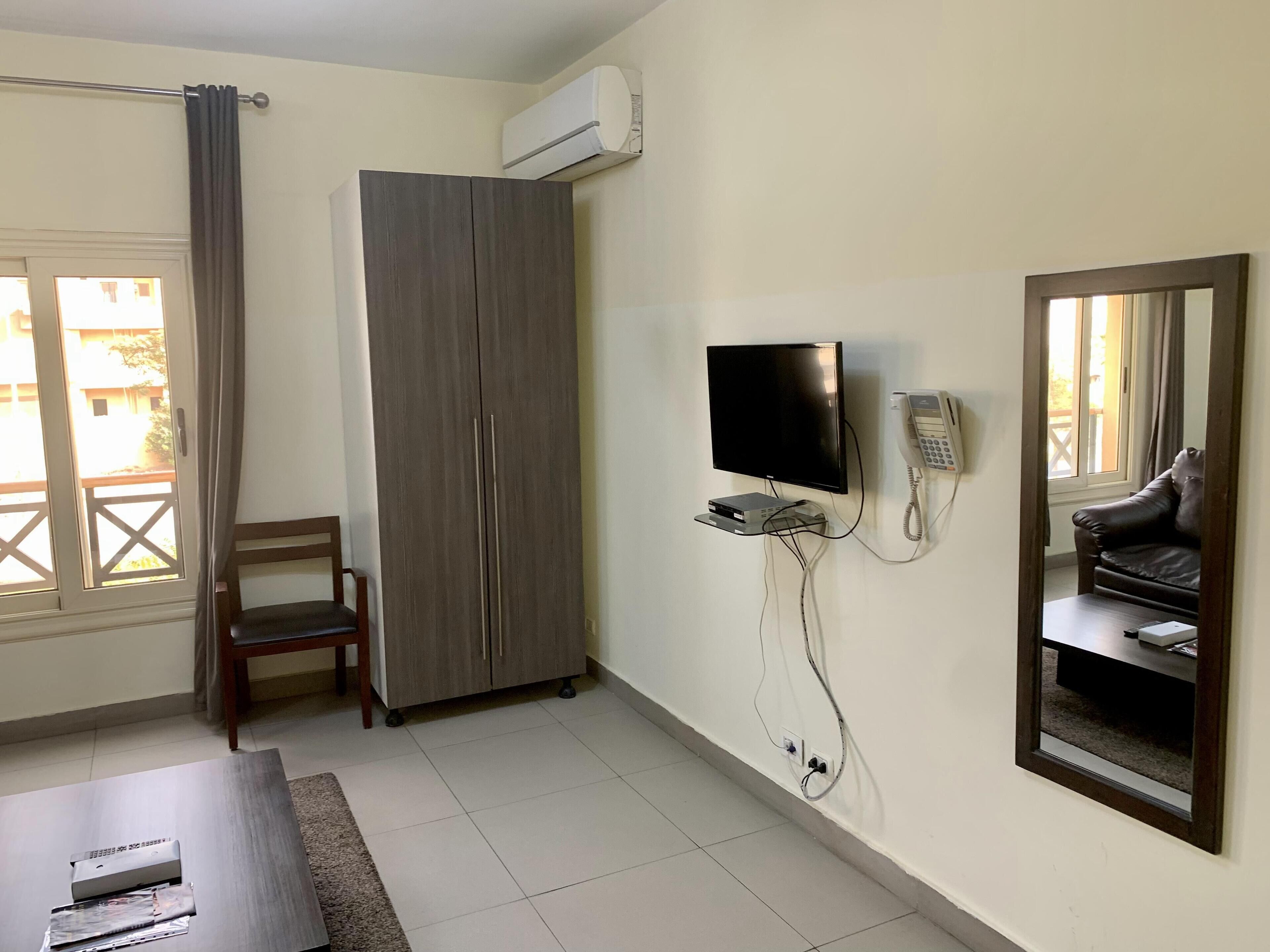 A room with a wardrobe, a television mounted on the wall, a telephone, and a window with a view of buildings outside.