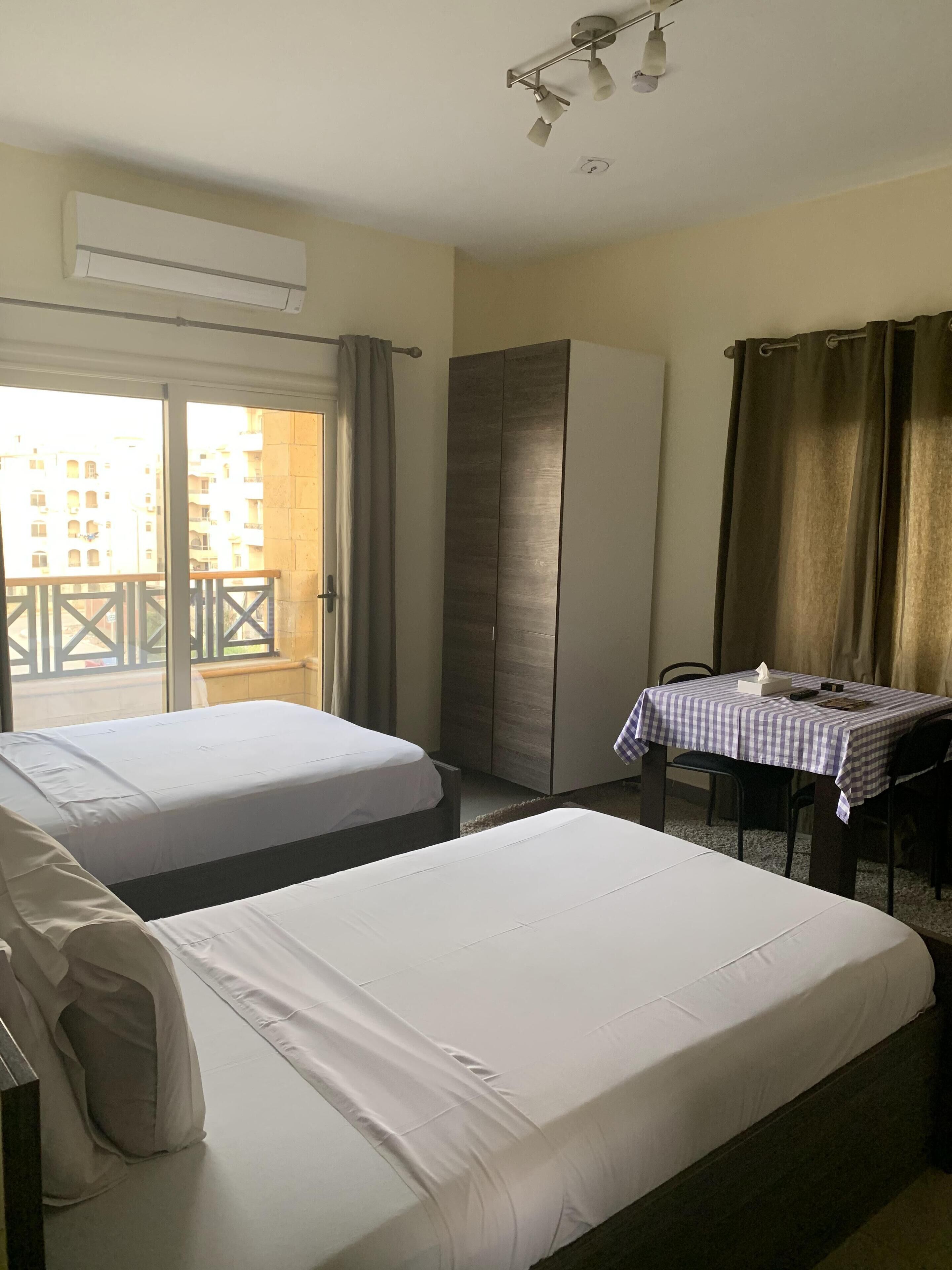 Habitación de hotel con dos camas, una mesa con mantel a cuadros, un armario y un balcón con vistas a edificios.