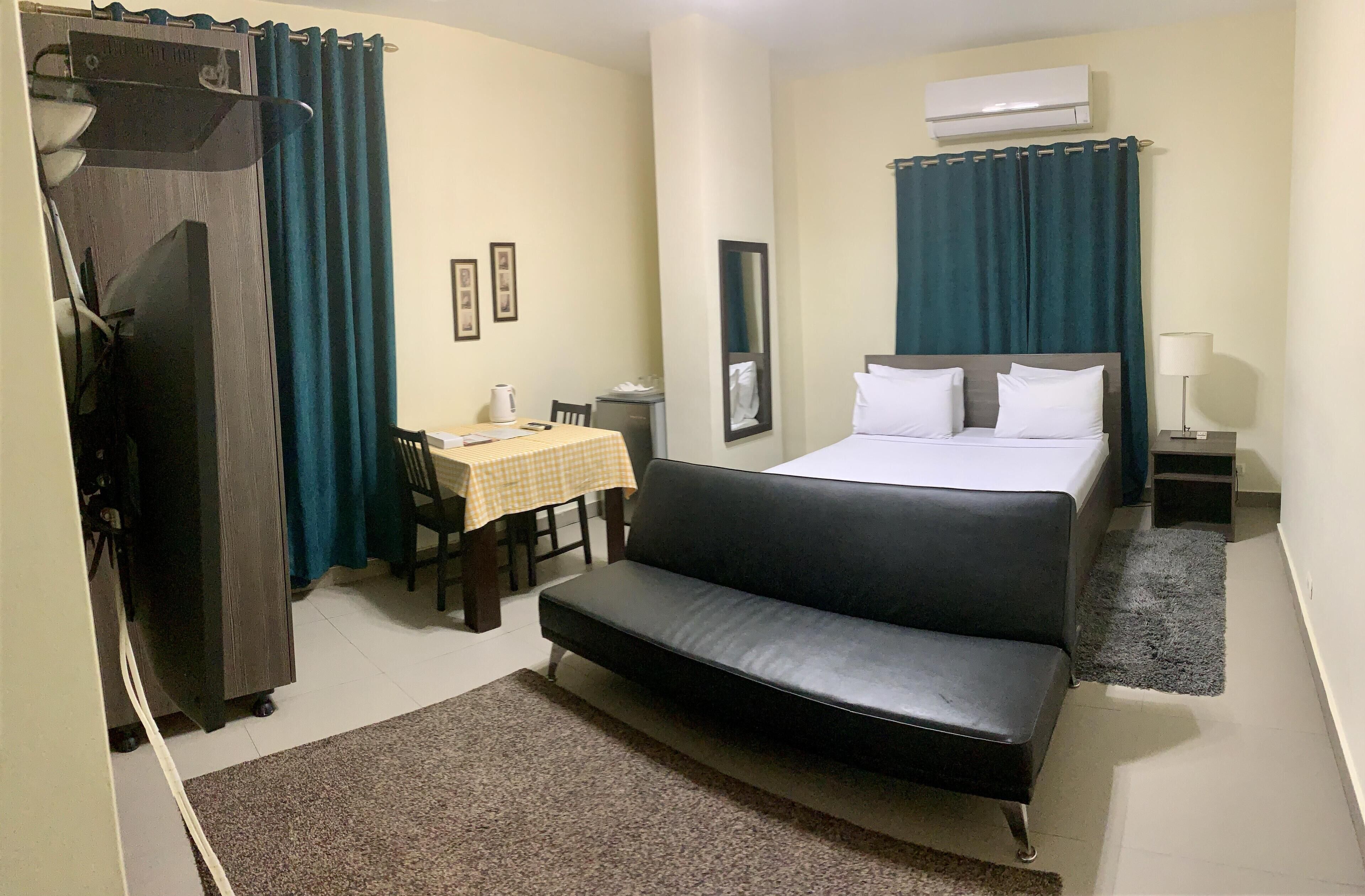 A hotel room with a bed, a sofa, a desk with a chair, a wardrobe, and an air conditioning unit.