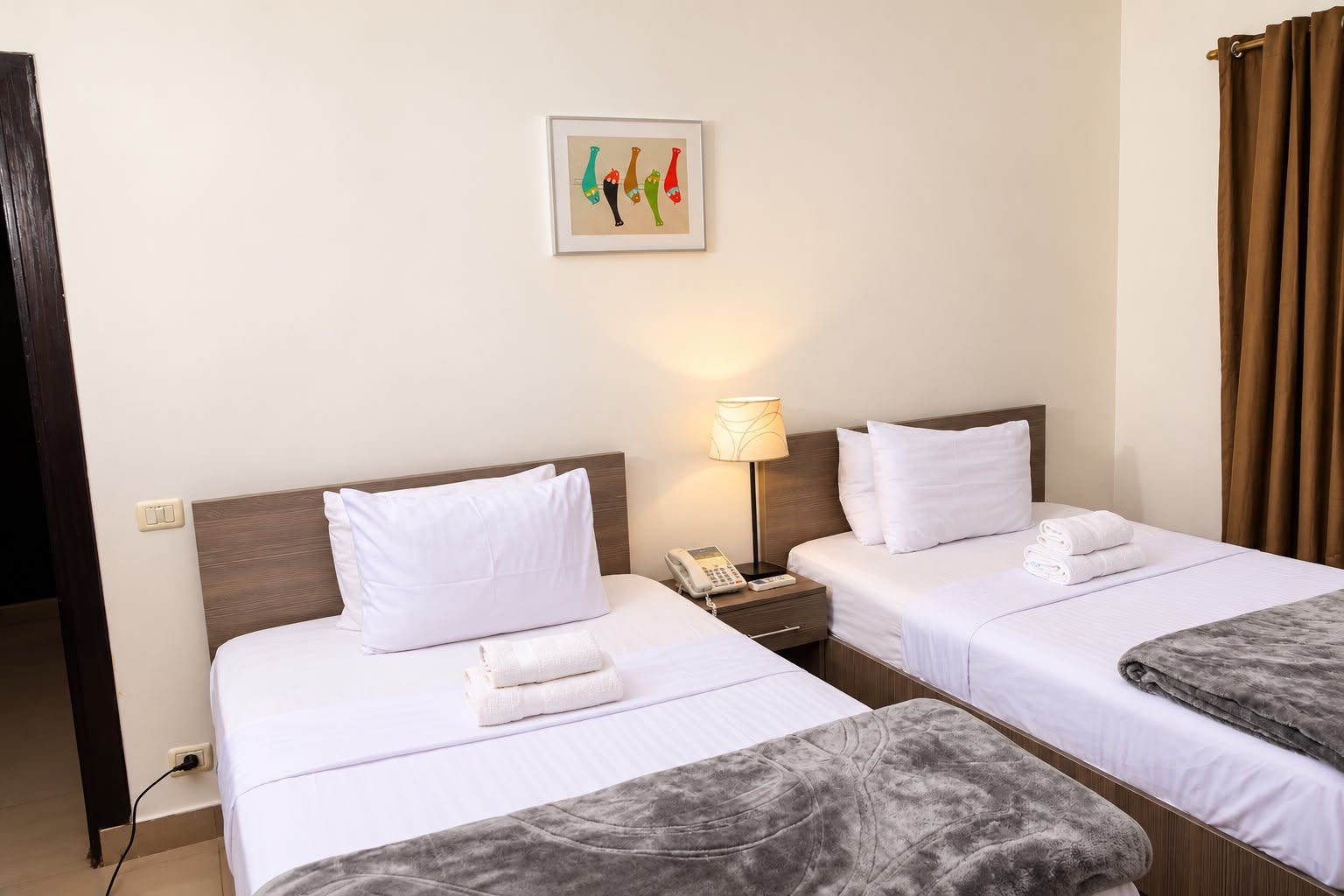 Hotellihuone, jossa on kaksi sänkyä, yöpöytä lampun kanssa, puhelin ja seinälle ripustettu taulu.