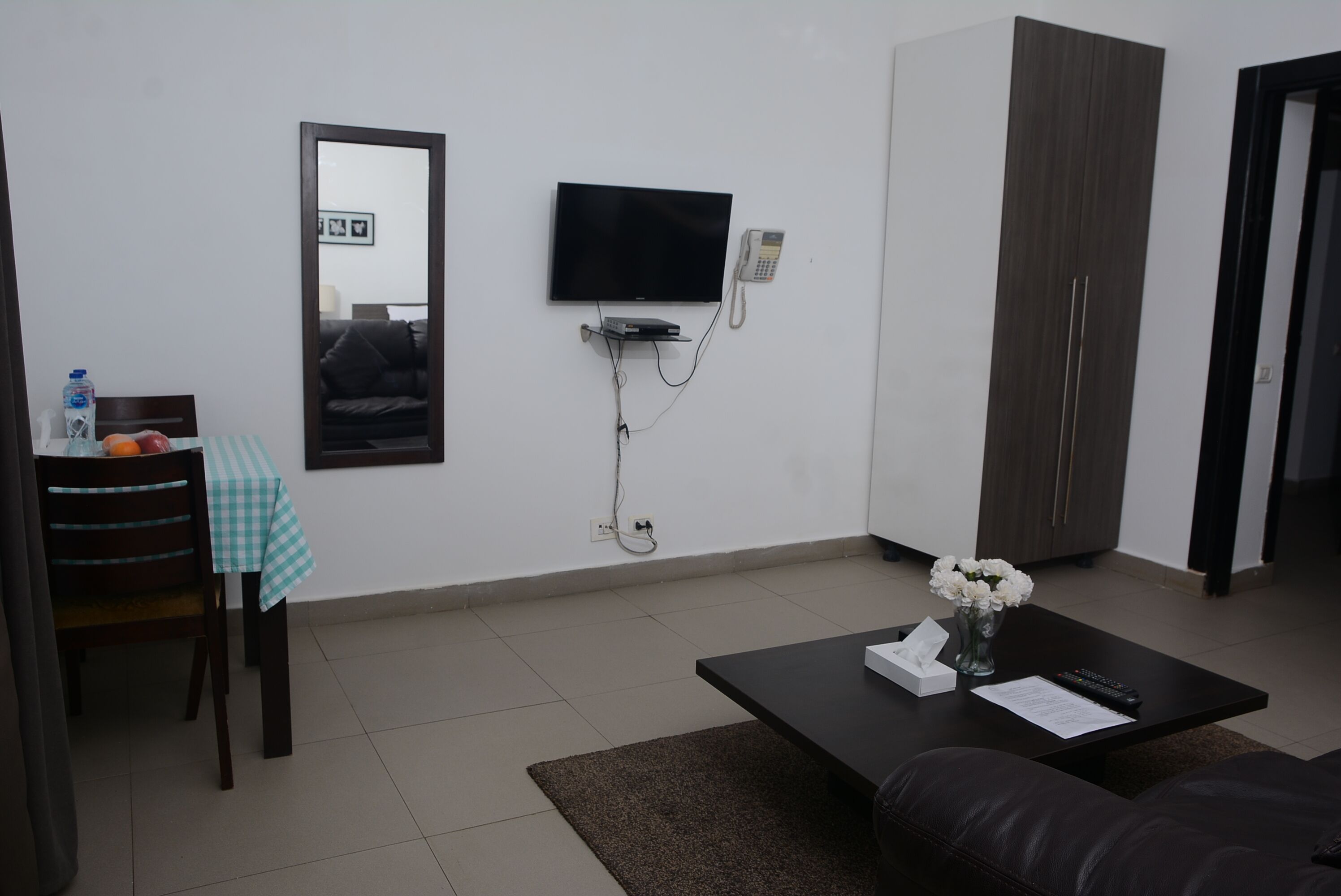 A modern living room with a flat-screen TV mounted on the wall, a small table with a checkered tablecloth, a coffee table with a vase of flowers, and a dark-colored sofa.