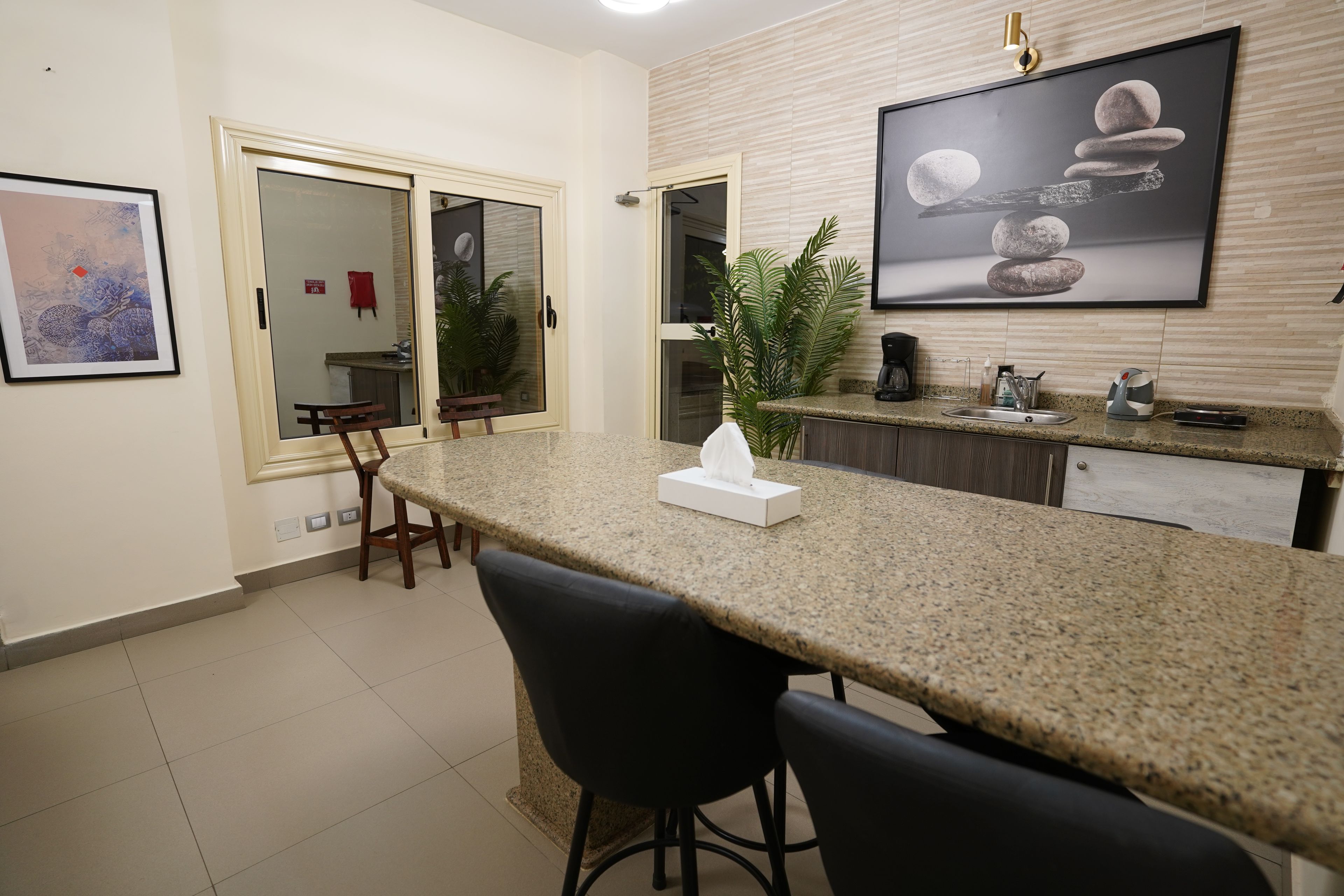 A modern kitchen with a granite countertop, bar stools, a coffee machine, and a framed artwork above the sink.