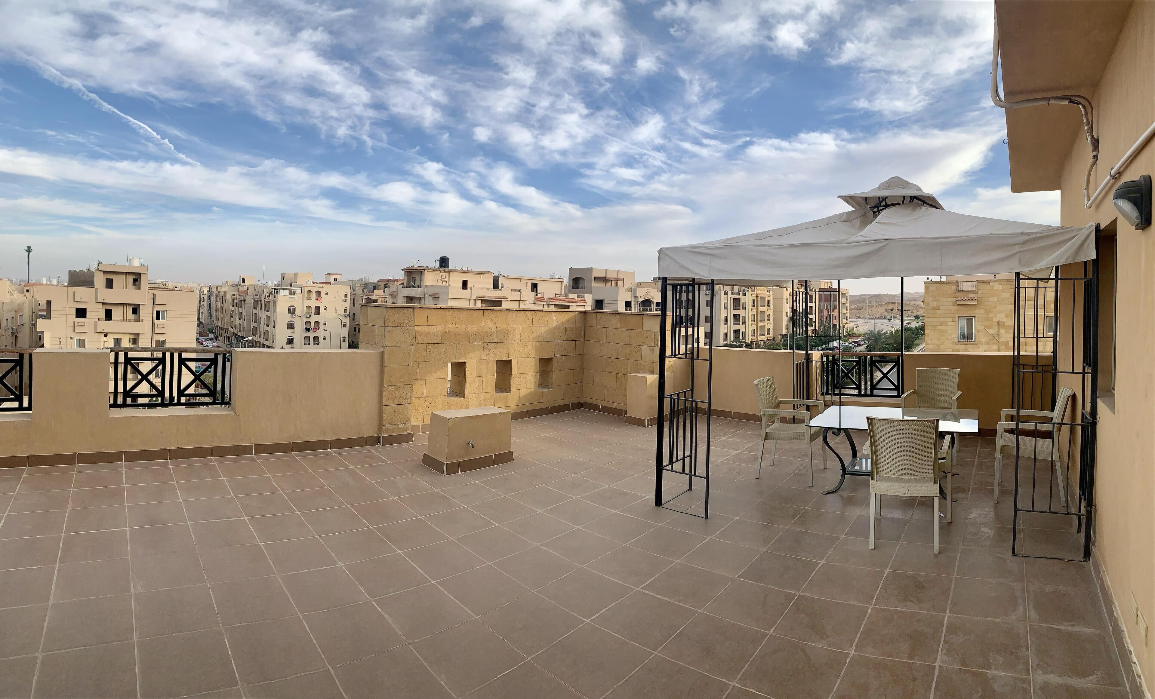 Una terraza en la azotea con suelo de baldosas, un espacio de estar cubierto con sillas y una mesa, y vistas de edificios circundantes.