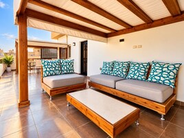 Un espacio al aire libre cubierto con sofás, una mesa de centro, vigas de madera y suelo de baldosas.