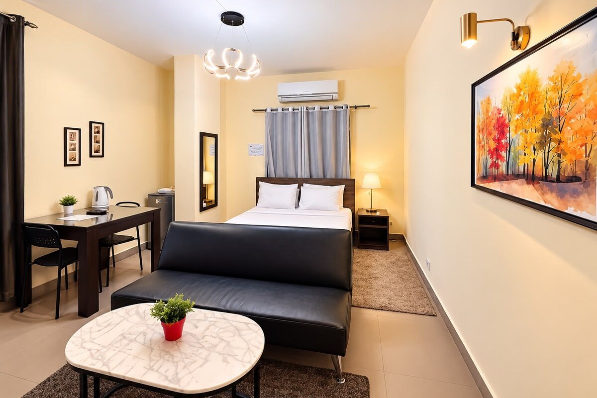 A hotel room with a bed, a sofa, a dining table, a small table with a plant, a painting of autumn trees, and a ceiling light fixture.