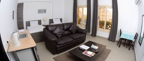 A modern hotel room with a sofa, a coffee table, a small kitchen area, and a balcony with curtains.