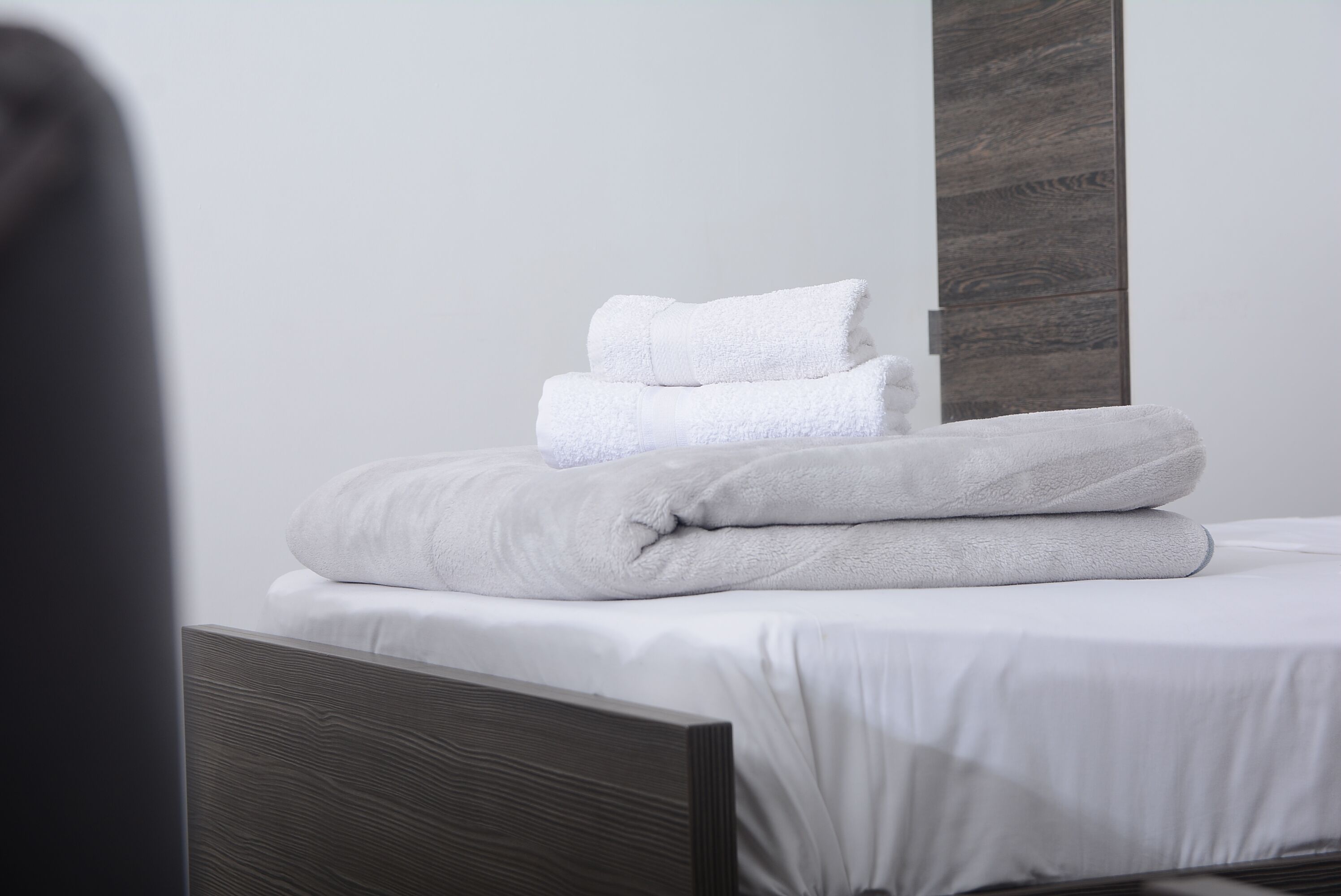 A neatly made bed with white linens and folded towels, a wooden headboard, and a plain wall with a vertical wooden panel.