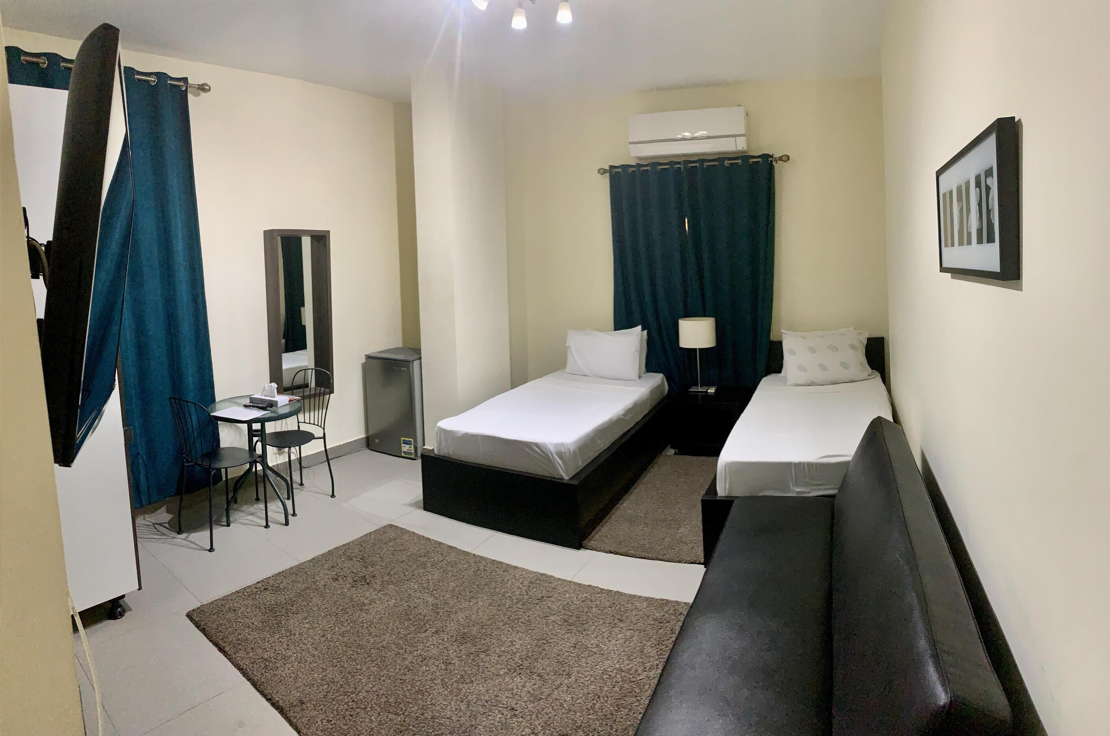 Ein Hotelzimmer mit zwei Einzelbetten, einem Schreibtisch mit Stuhl, einem kleinen Tisch, einem Spiegel und einer schwarzen Bank.