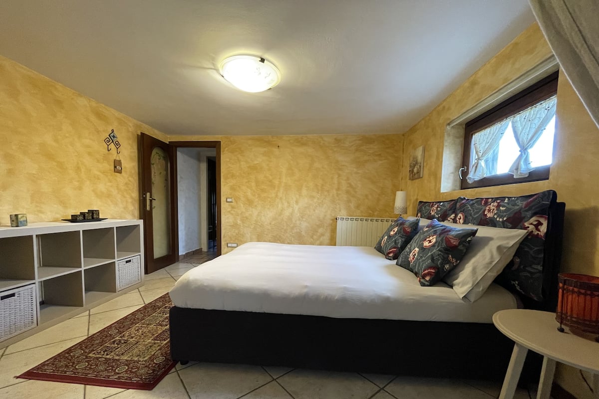 A bedroom with a bed, a bedside table, a window with curtains, a radiator, a shelf unit, and a door.