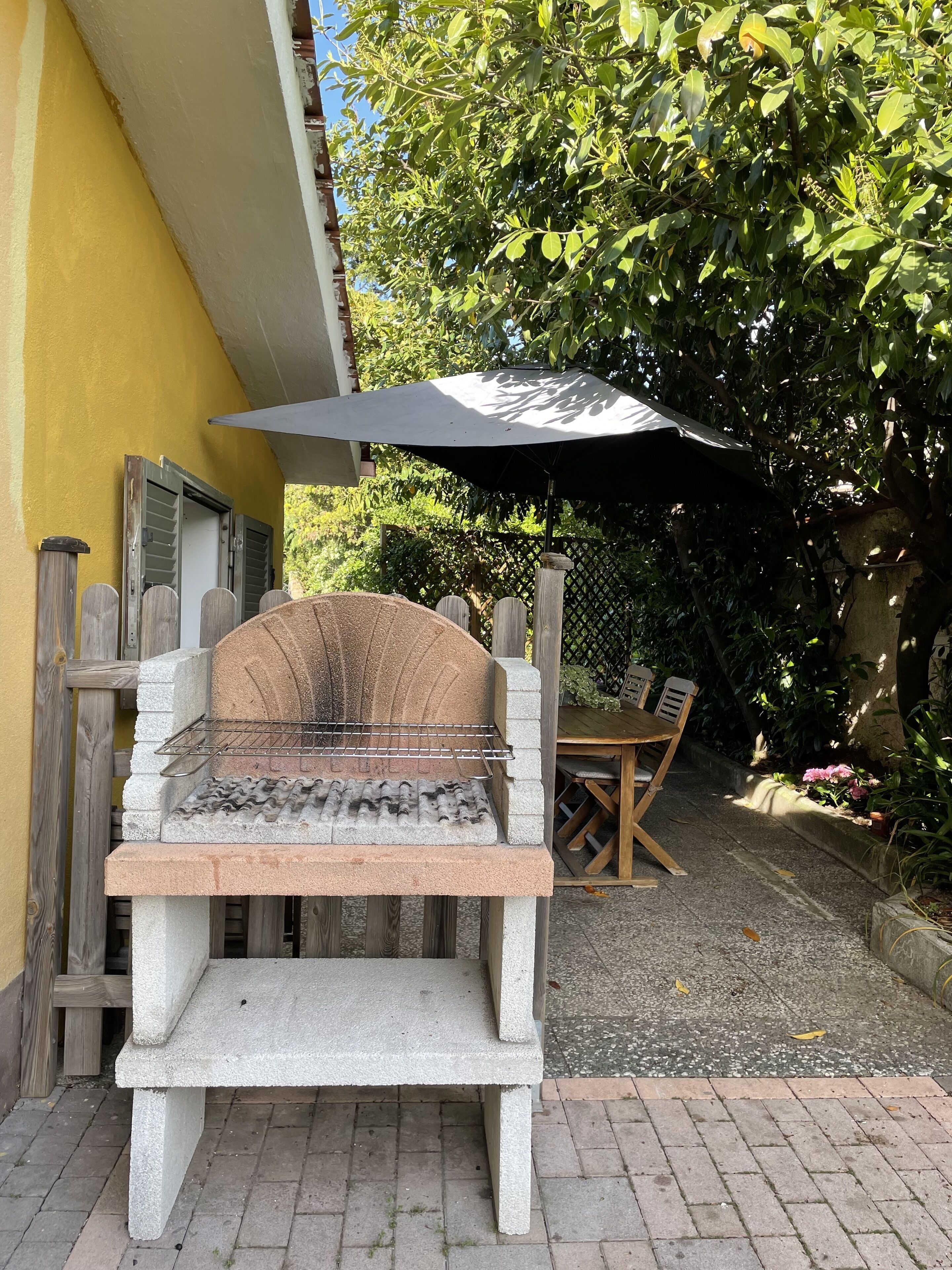 Un barbecue en béton avec une grille métallique, une table en bois avec des chaises et un grand parasol pour faire de l’ombre.