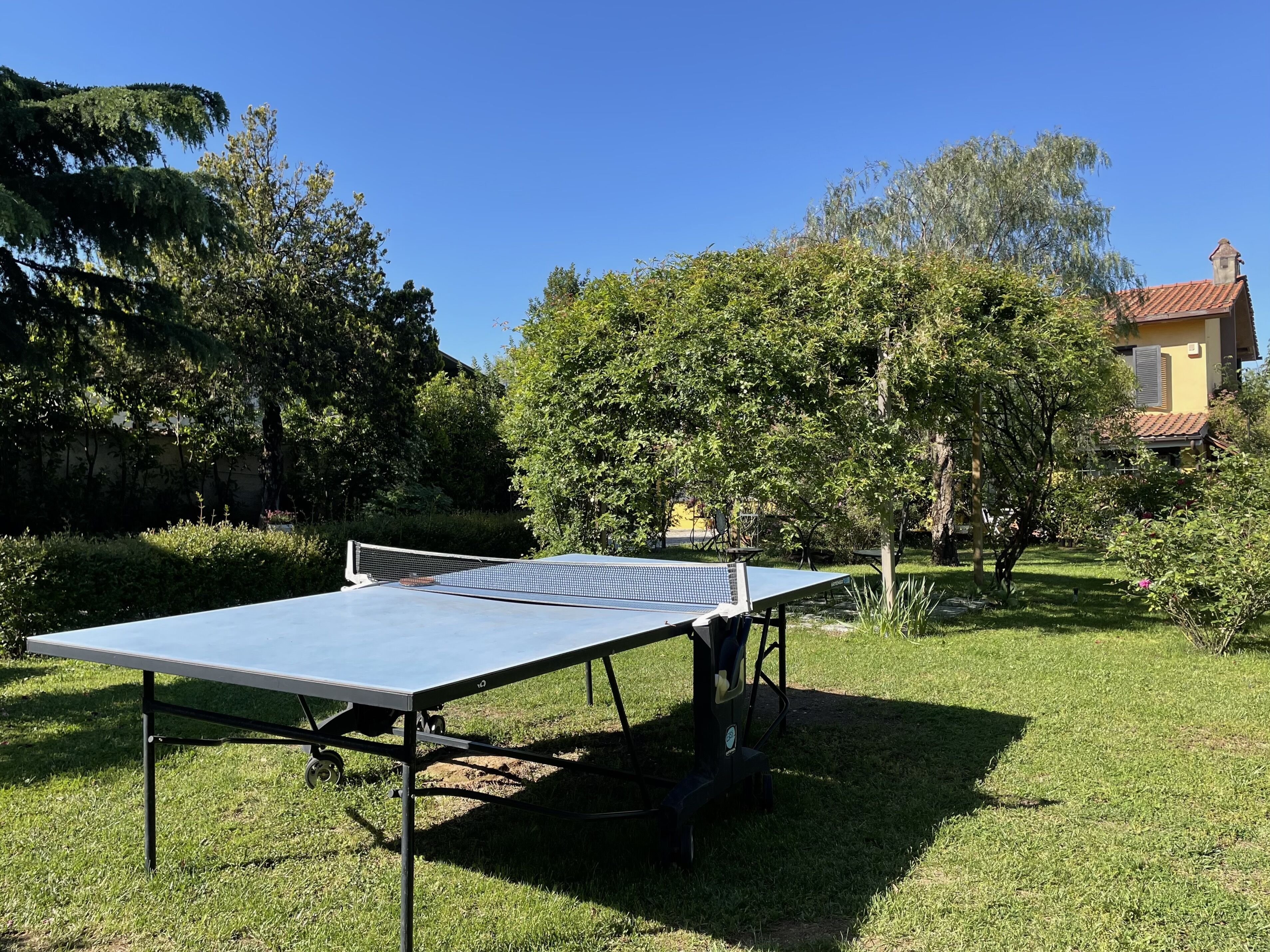 An outdoor ping pong table set up in a grassy area with trees and a building in the background.