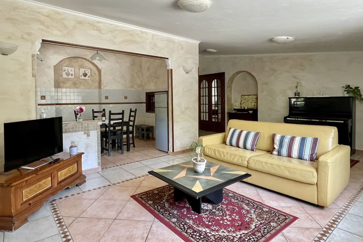 A living room with a TV, sofa, and coffee table. There is a kitchen in the background with a dining table and chairs.