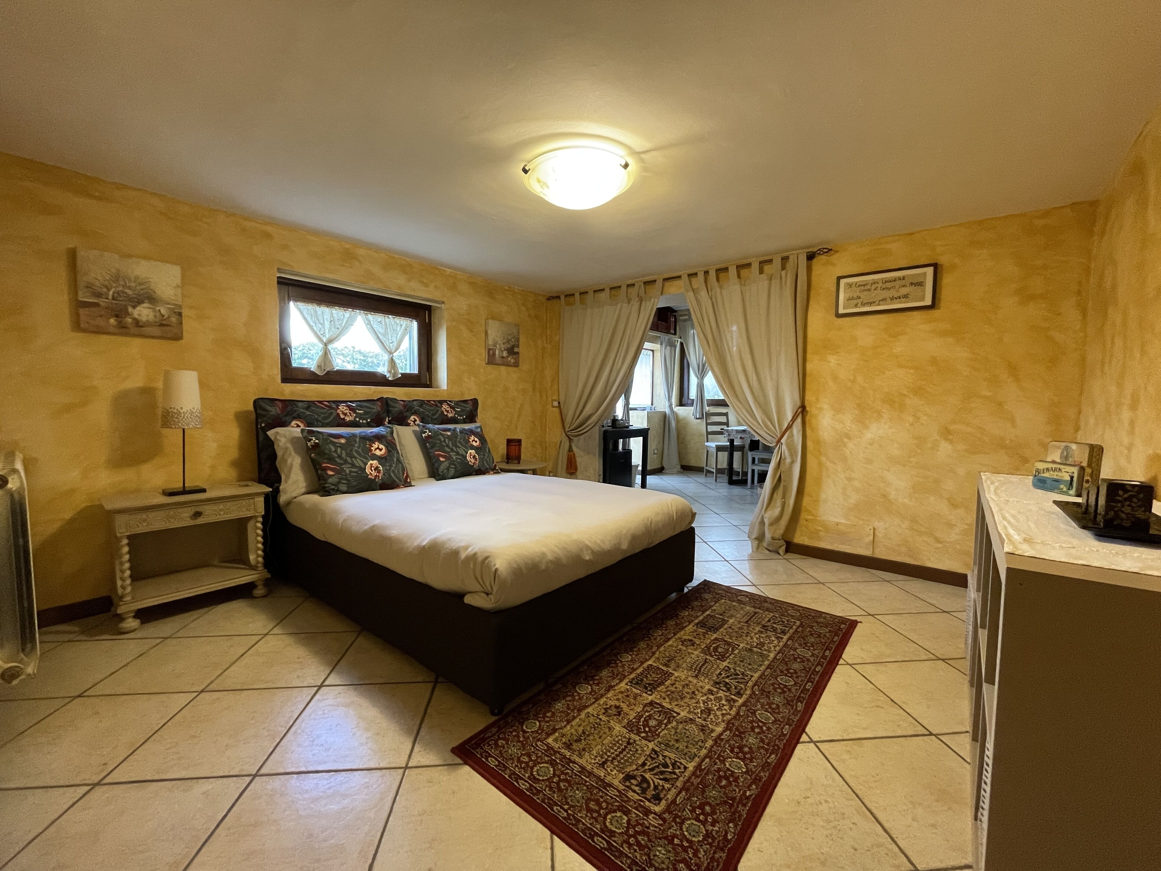 A bedroom with a bed, a nightstand, a desk, and a chair. There is a window with curtains, a ceiling light, and a rug on the floor.