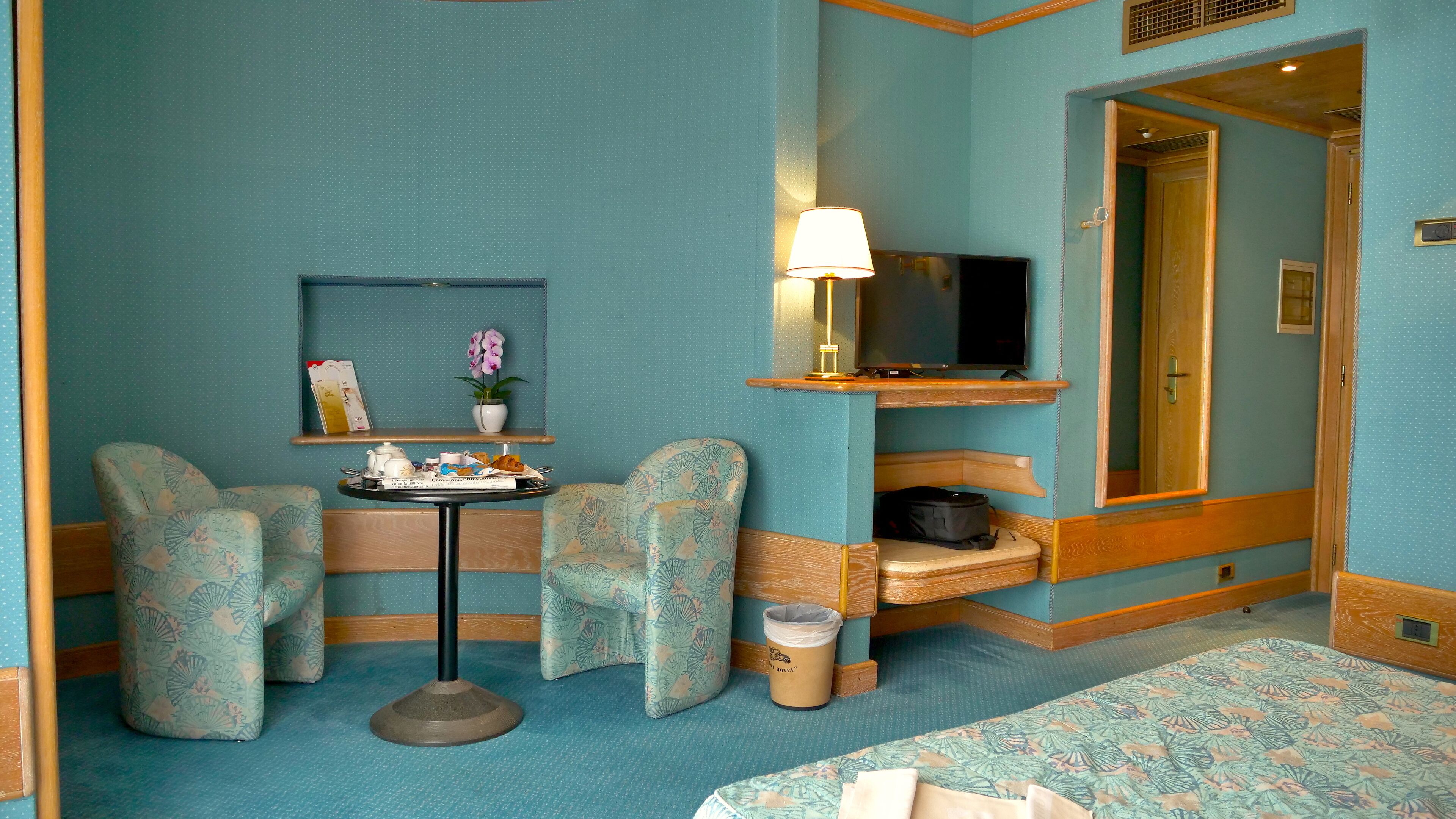 A hotel room with a bed, two chairs, a small table with a lamp, a television, and a mirror.