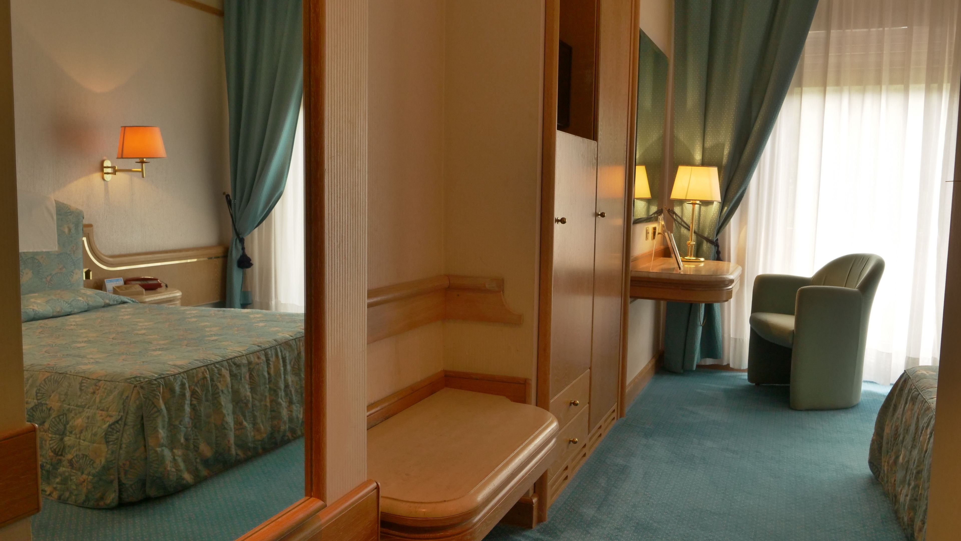 A hotel room with a bed, a bedside table, a chair, a desk with a lamp, and a mirror.