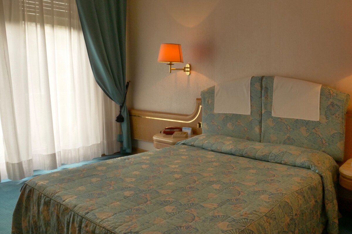 A hotel room with a bed, two bedside lamps, a nightstand, and a window with curtains.