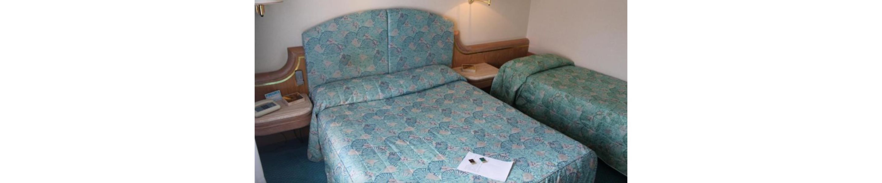 Una habitación pequeña con una cama individual, una colcha estampada, una mesita de noche con lámpara y un cajón.