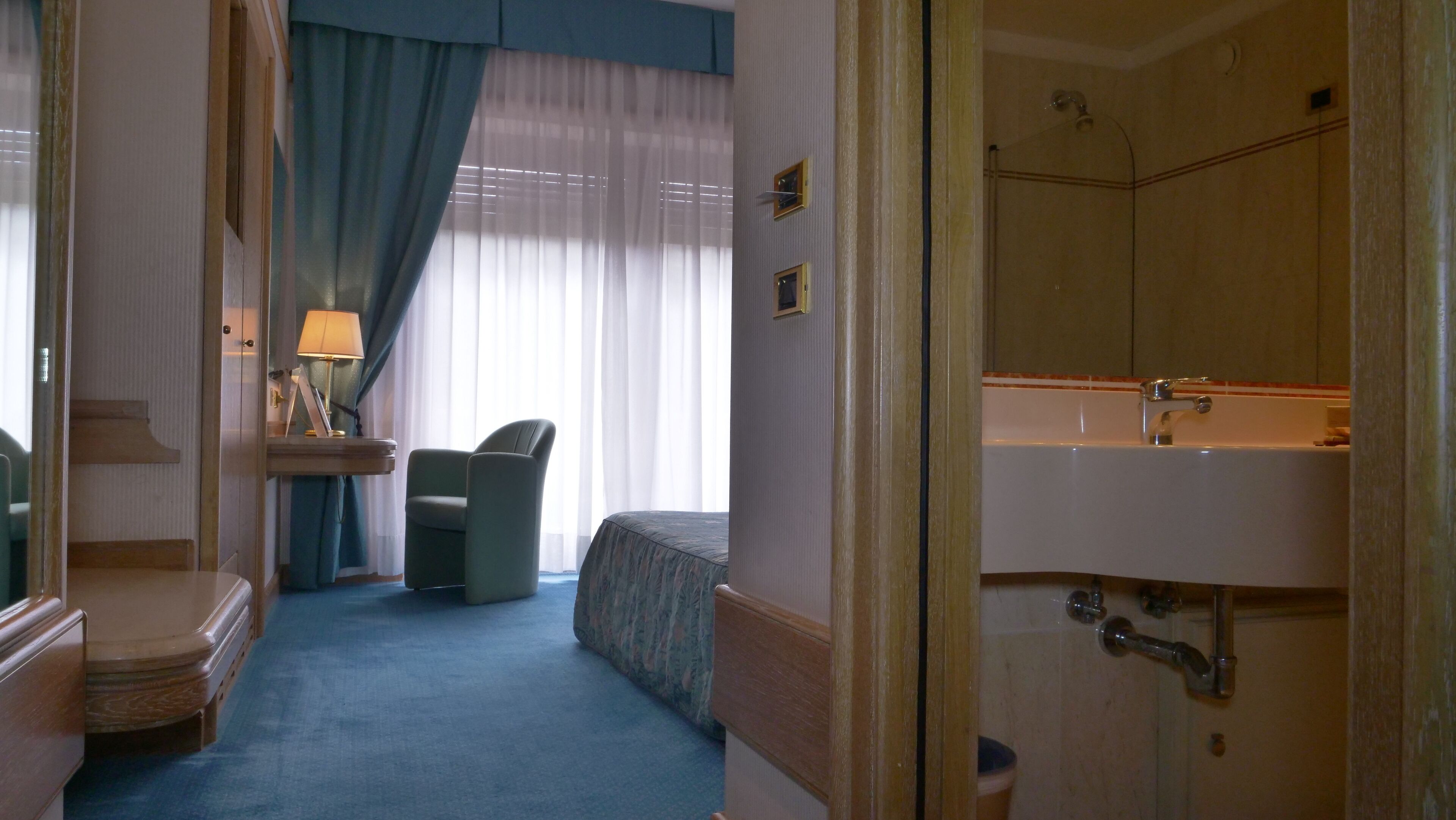 Habitación de hotel con cama, una silla, escritorio con lámpara y baño con lavamanos y espejo.