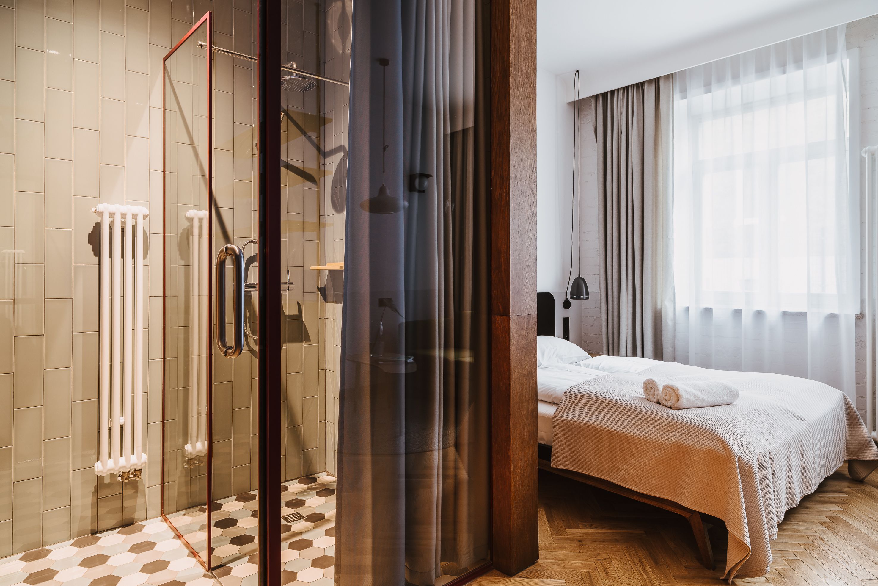 A modern hotel room with a glass-enclosed shower, a bed with a beige bedspread, and a large window with sheer curtains.