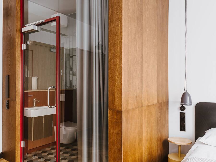 A modern hotel room with a wooden wardrobe, a glass shower enclosure, a bedside table with a lamp, and a bed with a white comforter.