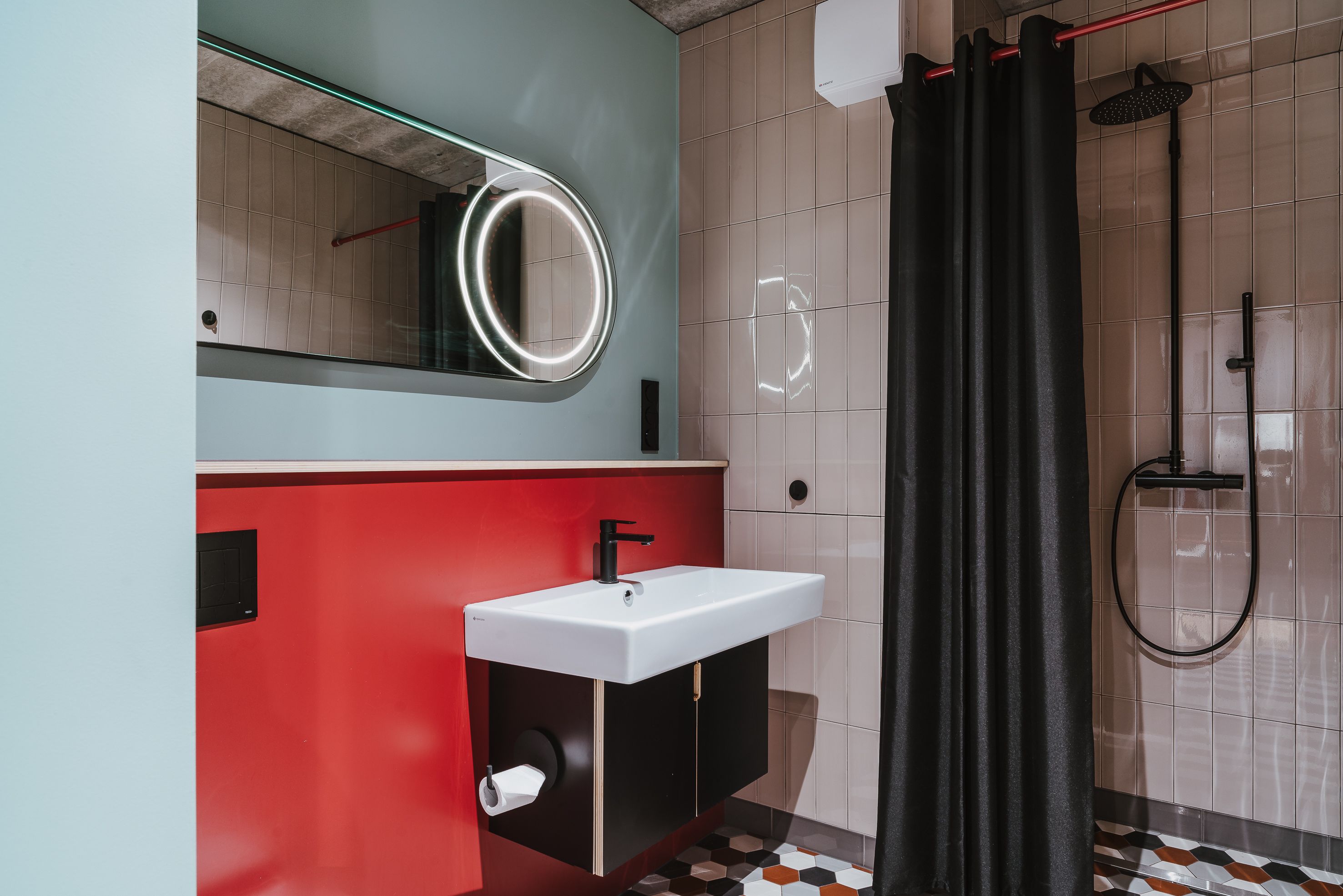 Un baño moderno con un tocador rojo, un lavabo blanco, un espejo con marco circular, una ducha con cortina negra y suelo de baldosas.