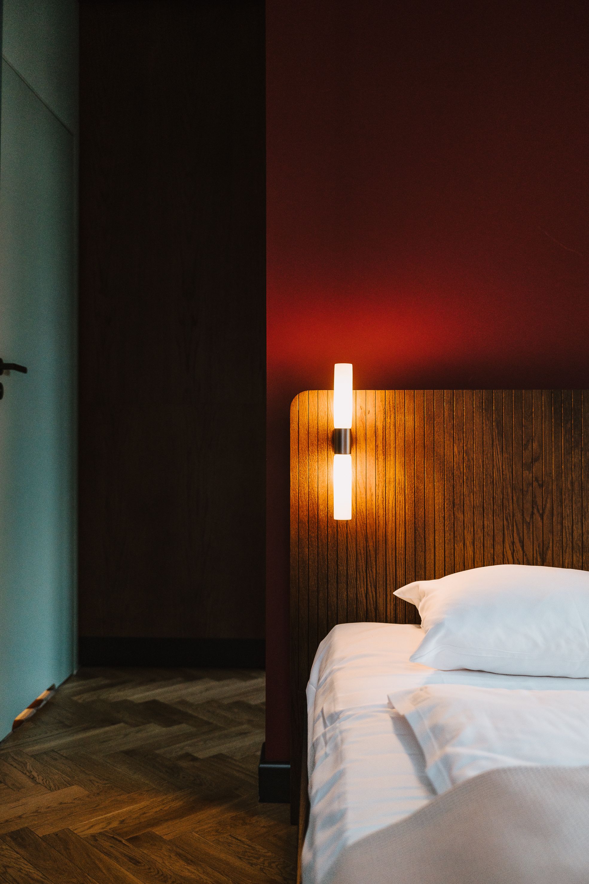 Una habitación de hotel con cabecera de madera, cama con ropa blanca y lámpara de pared.