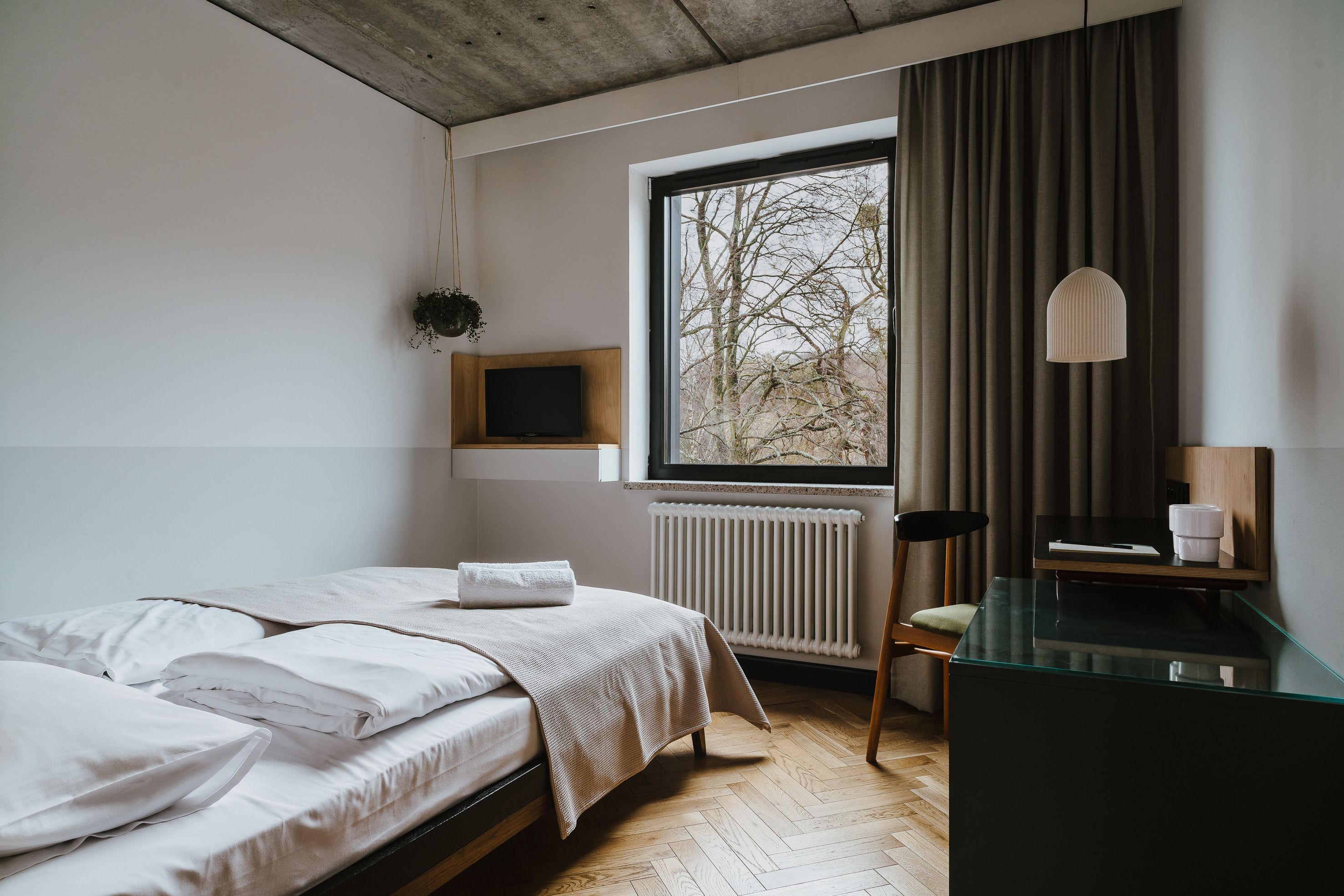 Een slaapkamer met een bed, bureau, stoel, raam, radiator en een hangplant.
