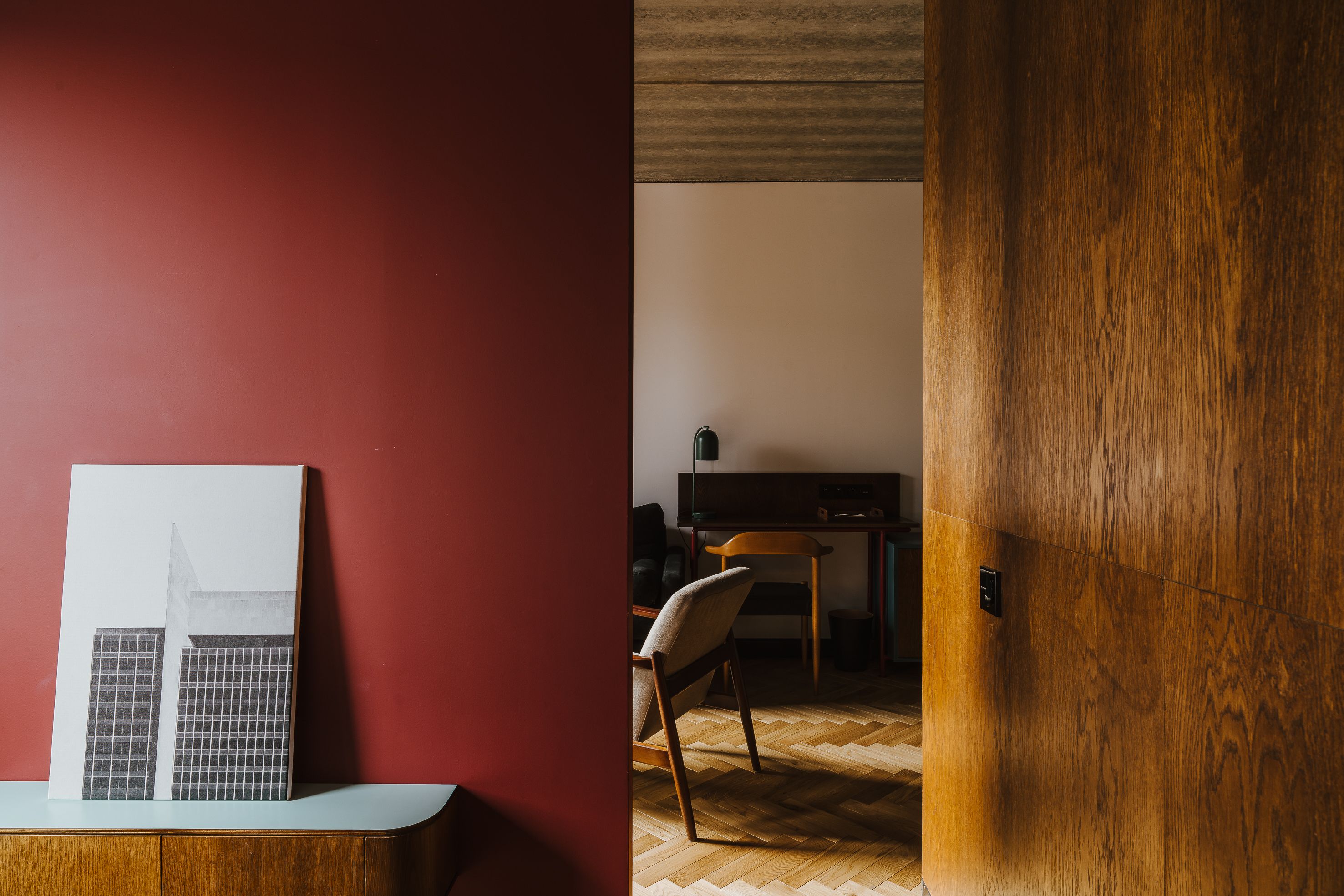 A room with a red wall, a wooden door, a white framed picture, a wooden chair, and a desk with a television.