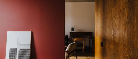 A room with a red wall, a wooden door, a white framed picture, a wooden chair, and a desk with a television.