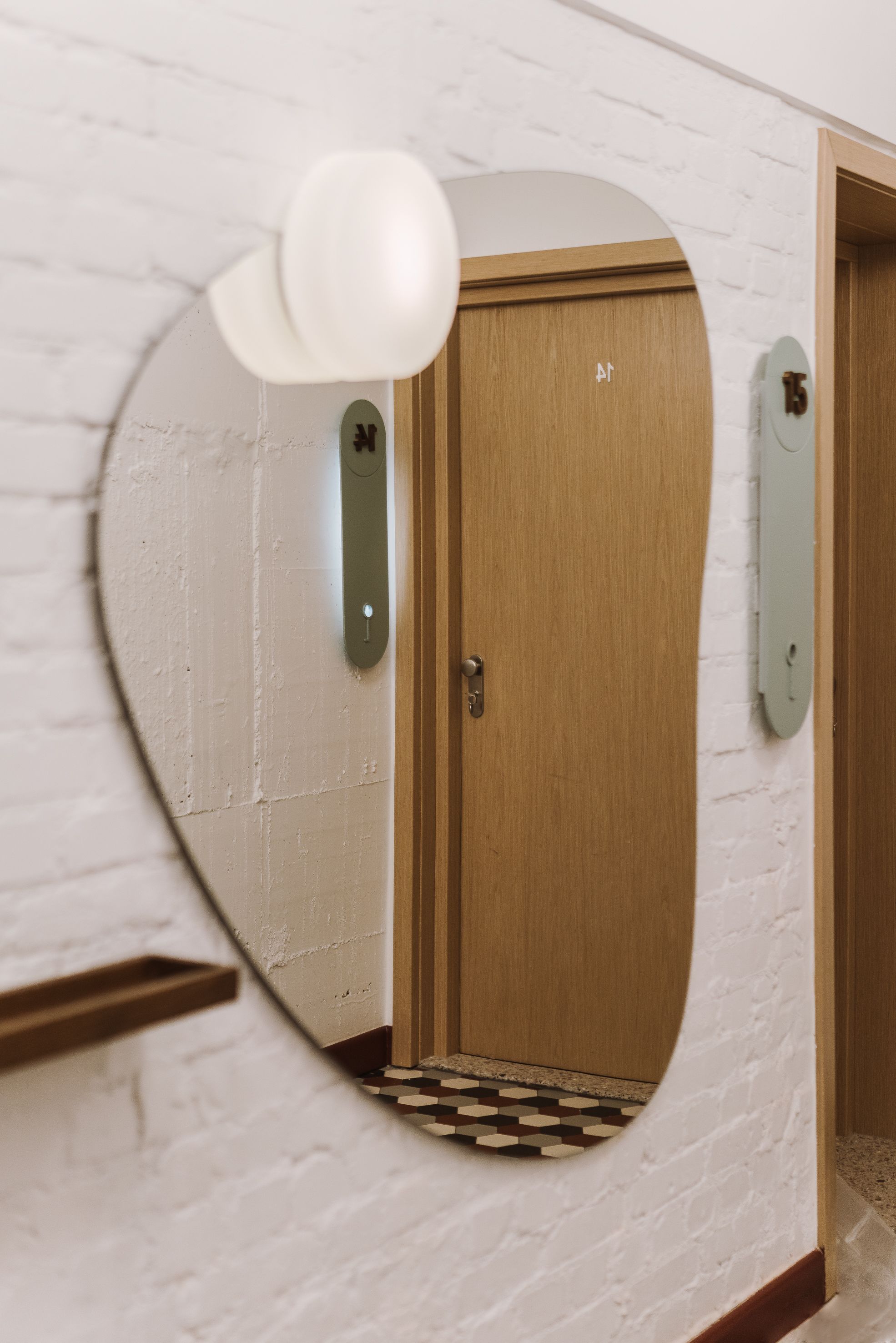 Un pasillo con un espejo redondo que refleja una puerta, una lámpara y una placa con un número.