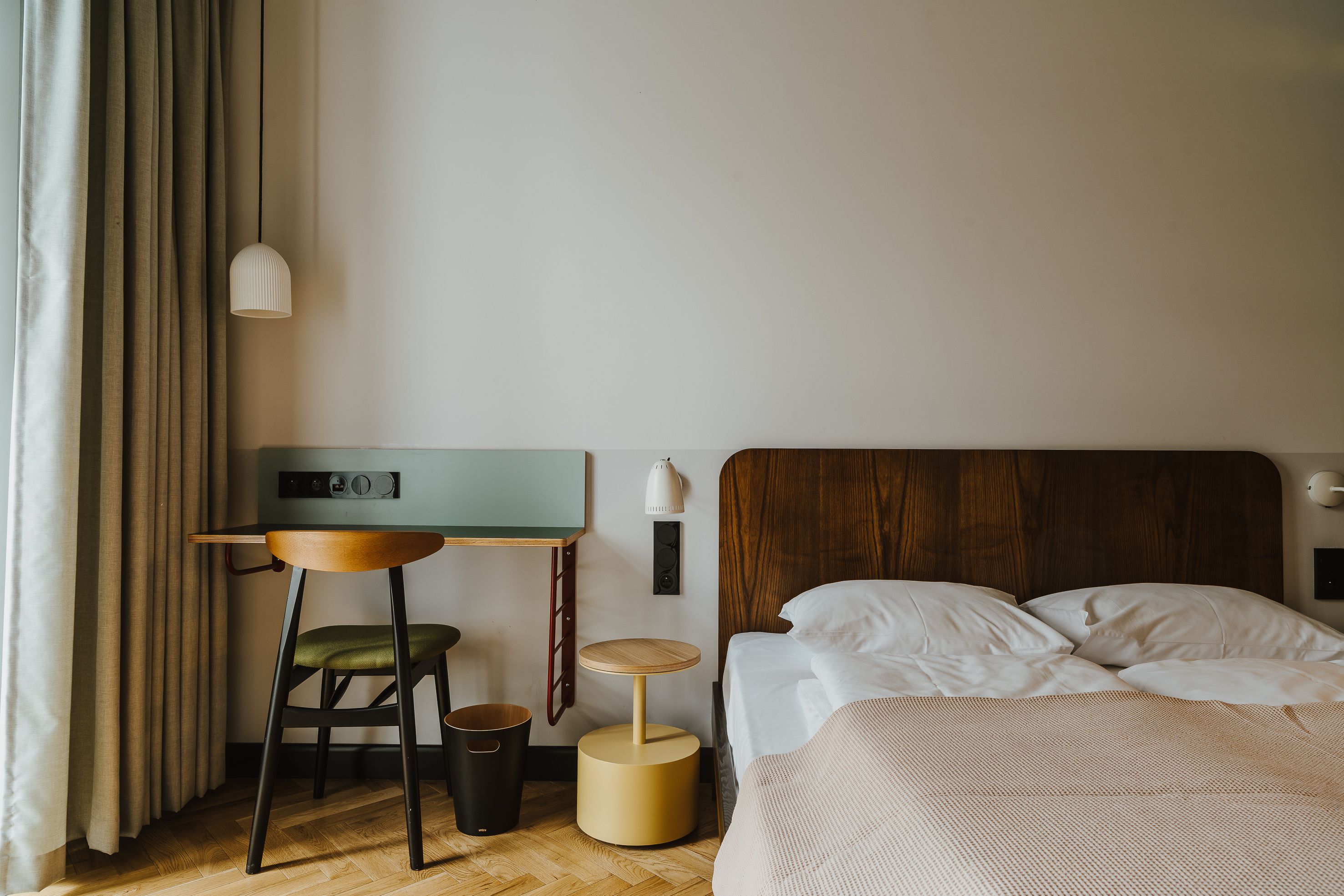 Un dormitorio con cabecera de madera, cama con ropa blanca, una mesita redonda con una silla verde, una lámpara de pared y suelo de madera.