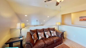 A cozy living room with a brown leather sofa, patterned cushions, a small side table with a lamp, and a ceiling fan.