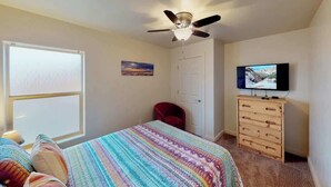 Un dormitorio con cama, ventilador de techo, televisor, cómoda y ventana.