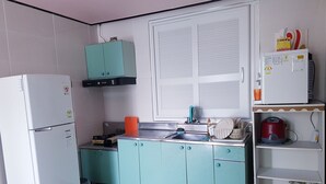 Eine kompakte Küche mit Kühlschrank, Herd und Spüle. Ein Fenster mit Jalousien, ein türkisblaues Schränkchen und eine Mikrowelle auf einem Regal.