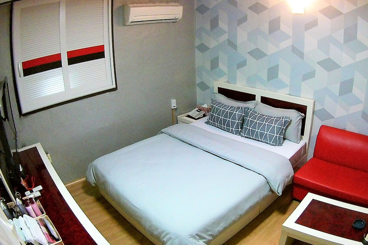 A bedroom with a bed, a red chair, a bedside table, a nightstand, a wall-mounted air conditioner, and a geometric-patterned wall.