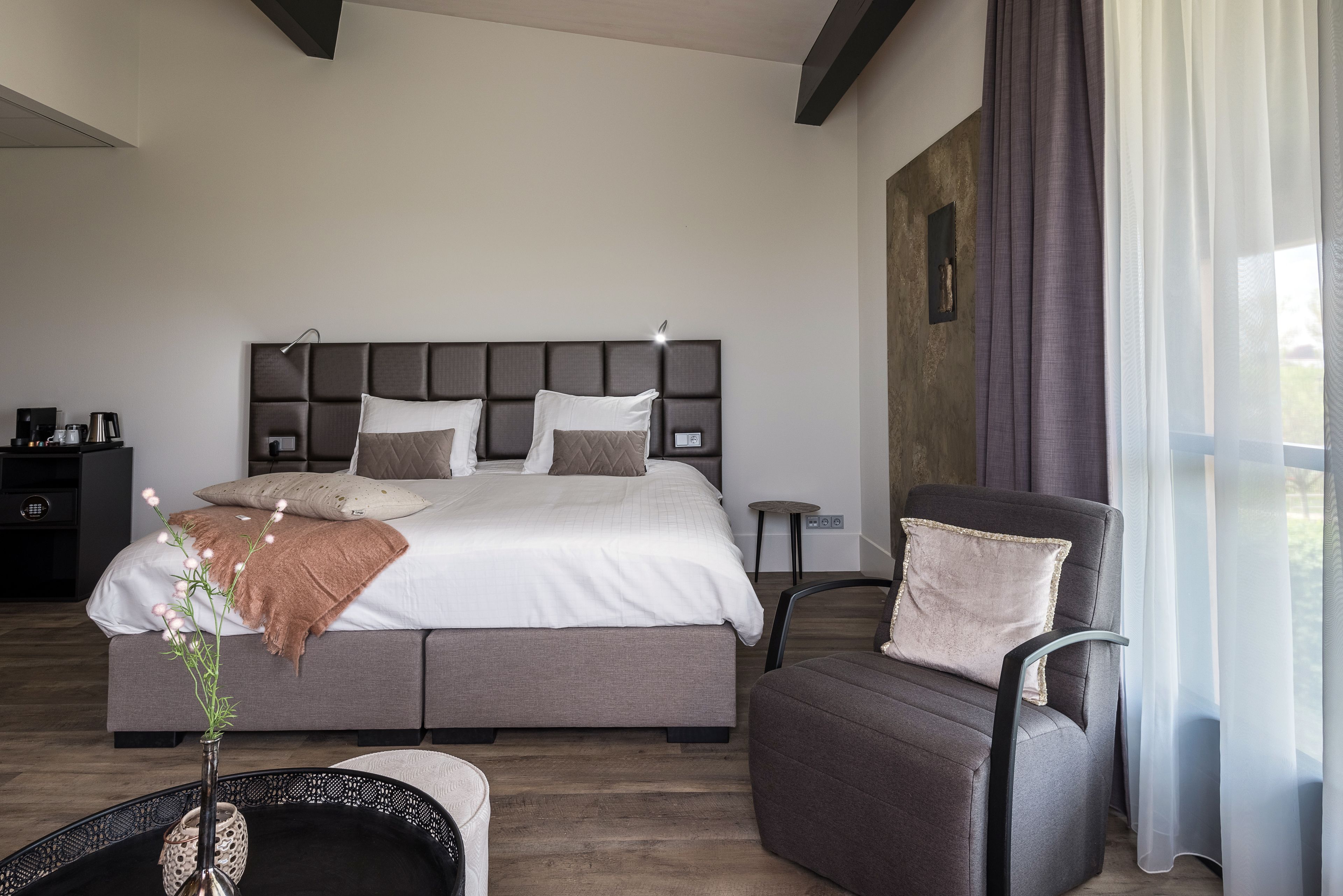 A modern bedroom with a large bed, a side table, a chair, and a window with curtains.