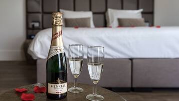 Una bottiglia di champagne Henkell Trocken, due calici e petali di rosa su un tavolo rotondo in una camera d'albergo, con un letto e una testiera sullo sfondo.