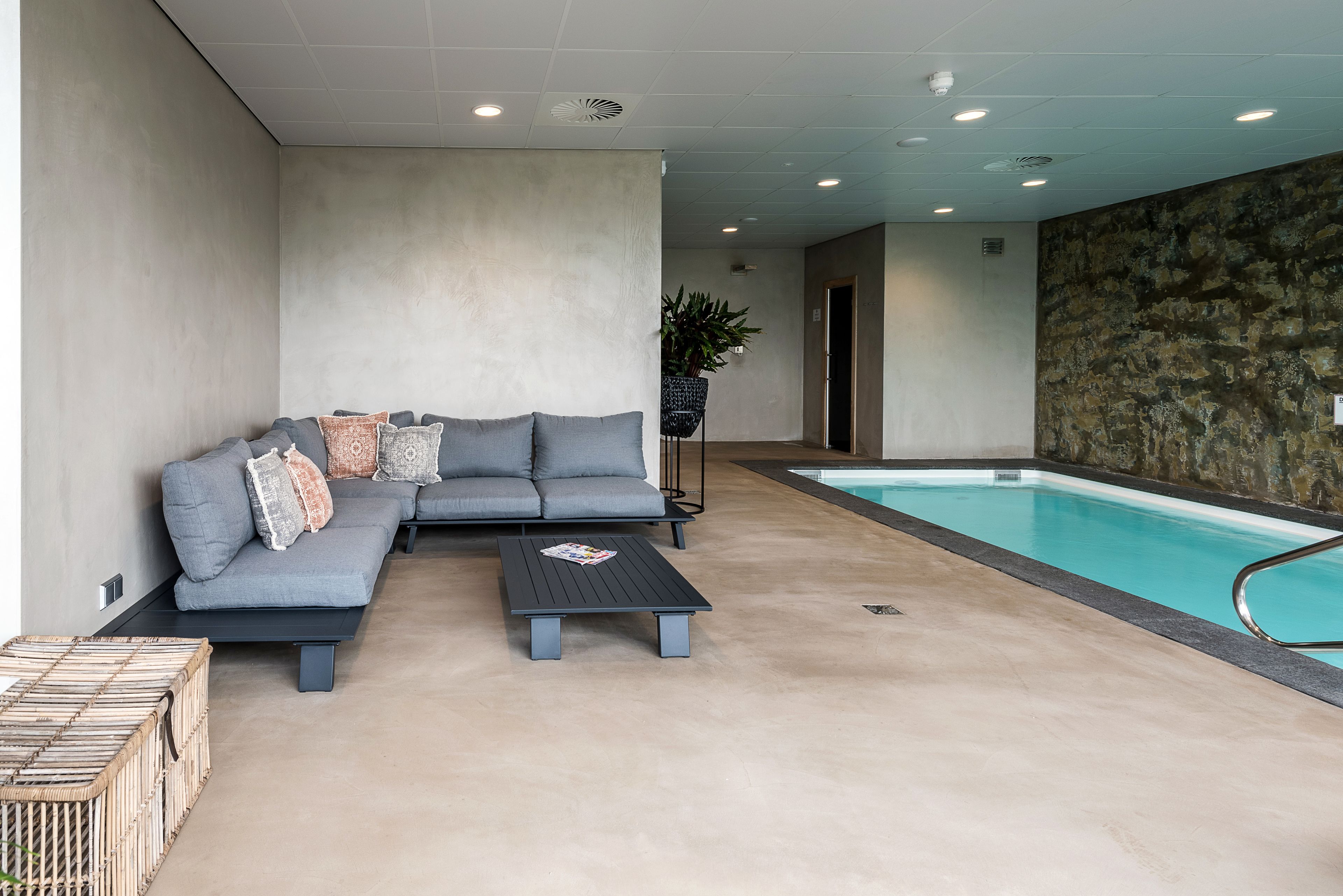 A modern indoor pool area with a grey sectional sofa, a small table, and a wicker basket.
