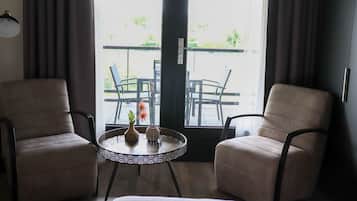 A room with two beige armchairs, a small round table with a vase and a decorative item, and a view of an outdoor patio with chairs and tables.