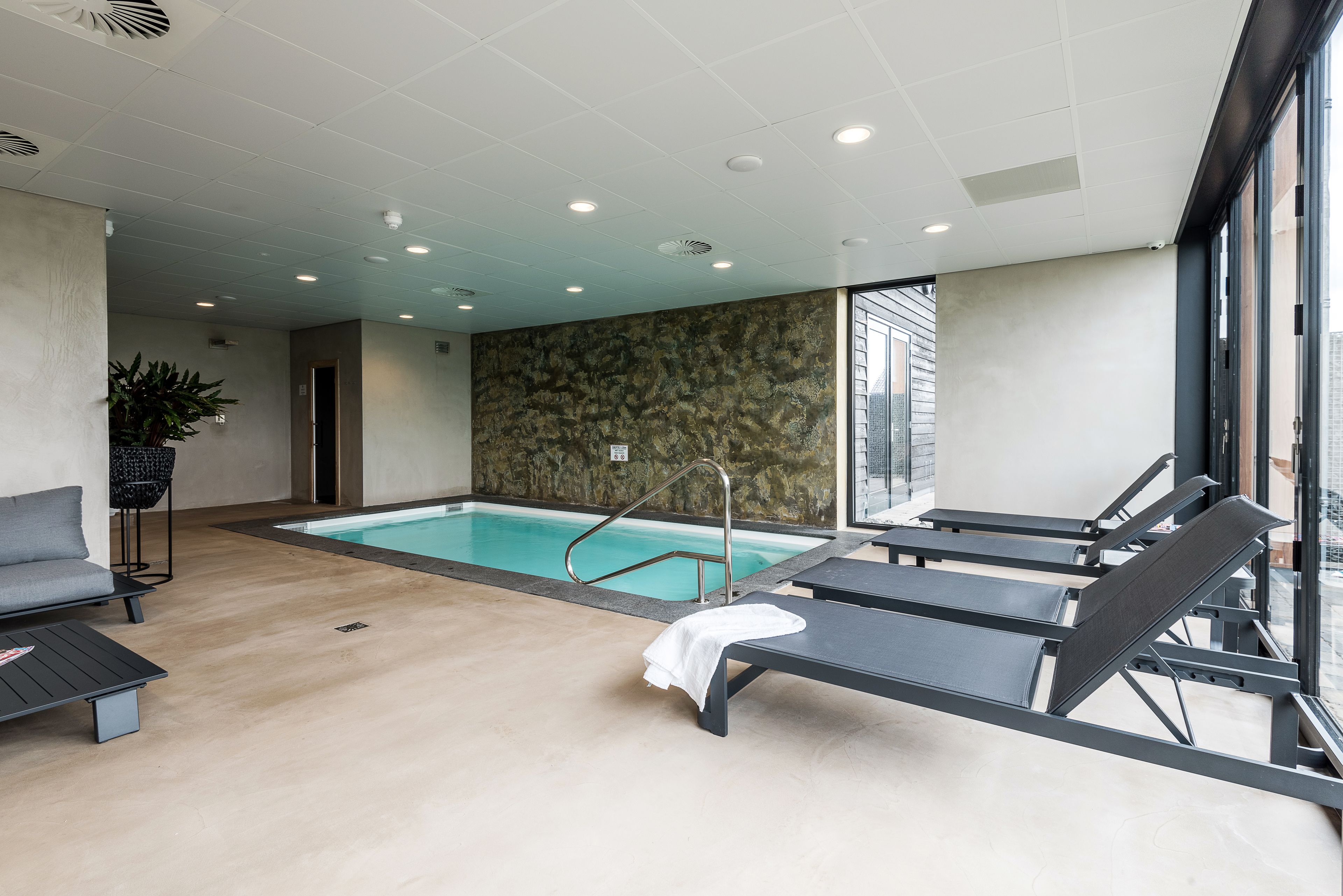 A modern indoor pool area with lounge chairs, a small seating area, and a stone wall.