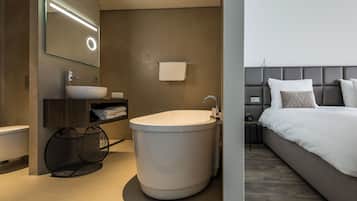 A modern bathroom with a freestanding bathtub, a large mirror, and a bedside table with a lamp.