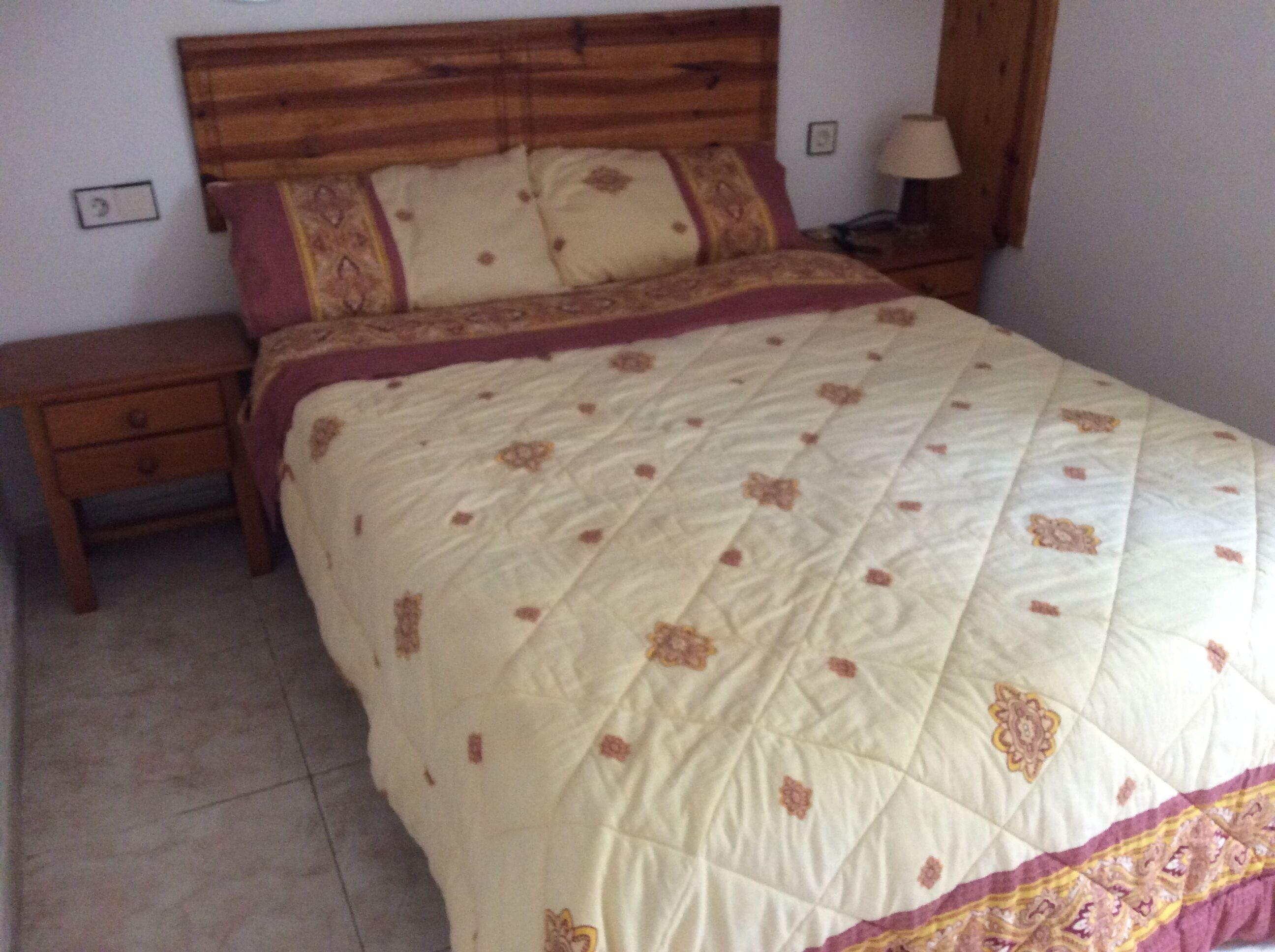 A bed with a quilted cover, a wooden headboard, and a nightstand with a lamp.