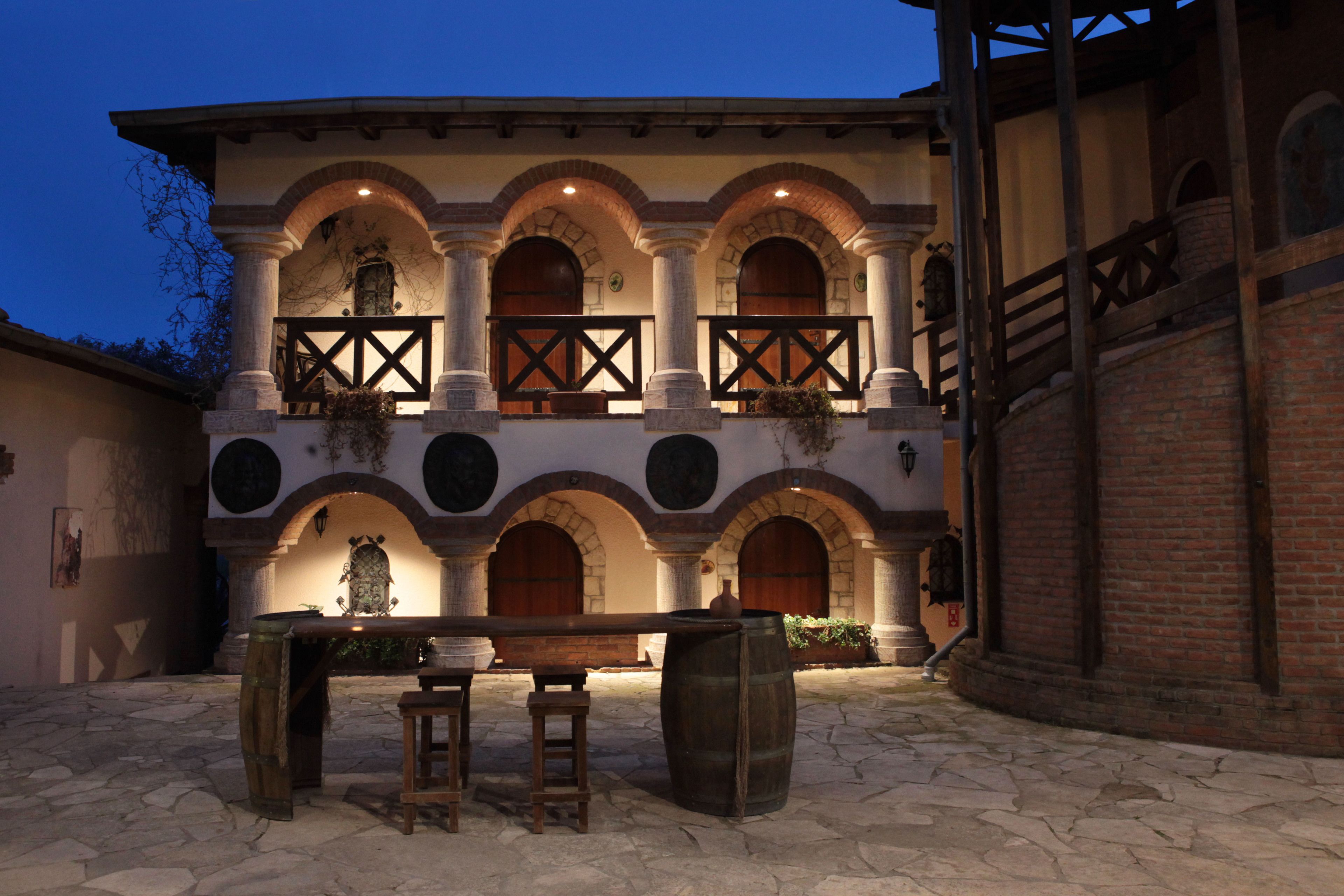 Un patio con piso de piedra, un banco de madera y una mesa. Hay dos taburetes de madera y un barril grande. El edificio tiene fachada de ladrillo con arcos y un balcón con barandilla de madera.