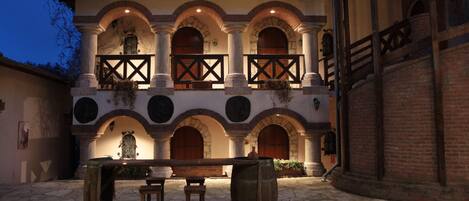 Un patio con piso de piedra, un banco de madera y una mesa. Hay dos taburetes de madera y un barril grande. El edificio tiene fachada de ladrillo con arcos y un balcón con barandilla de madera.