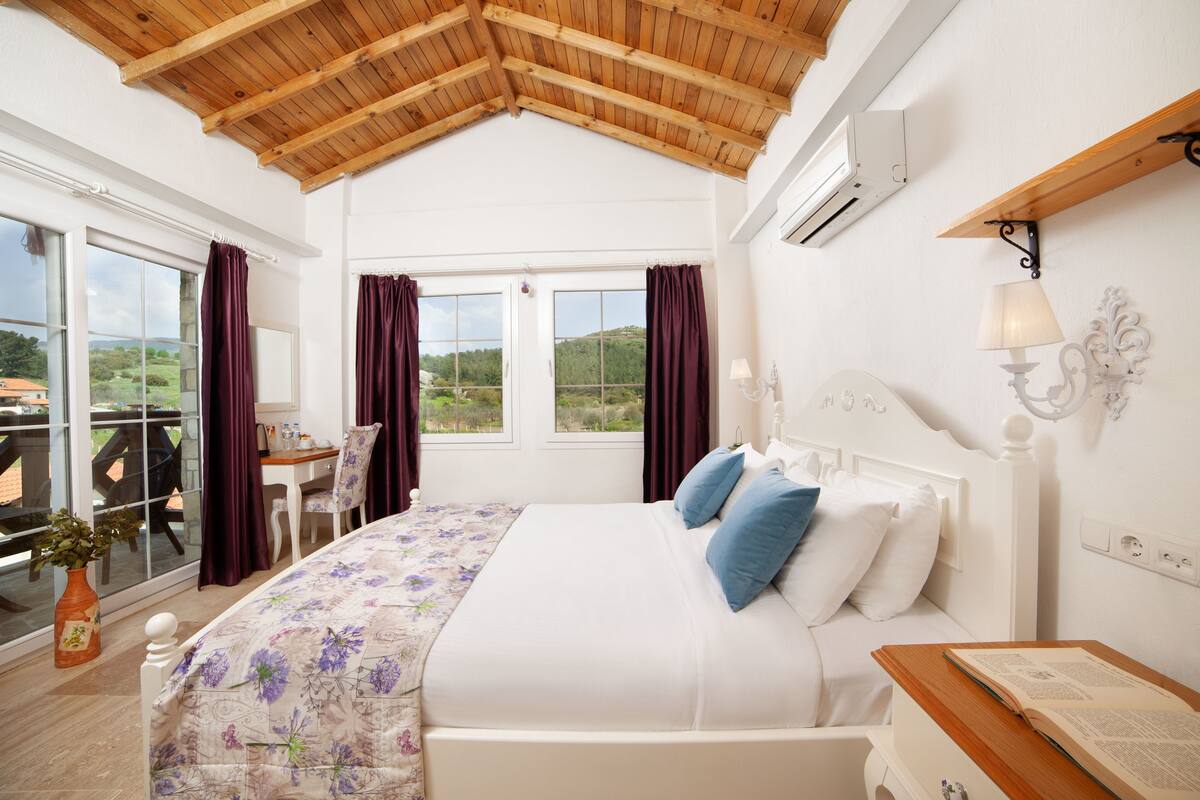 A bedroom with a wooden ceiling, a bed with a floral bedspread, a desk with a chair, and a view of the outdoors.