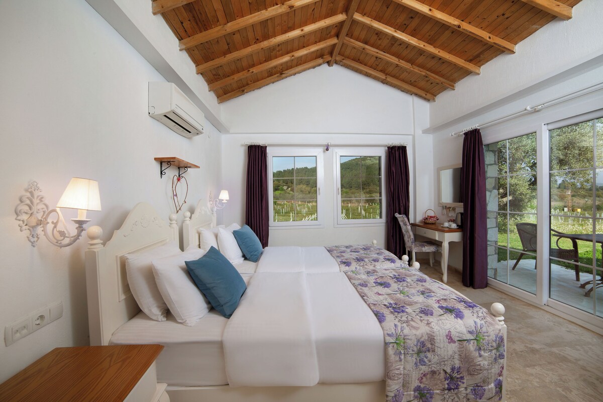 Une chambre avec un plafond en bois, un lit recouvert d’une couvre-lit à motifs floraux, un bureau et une vue sur l’extérieur.